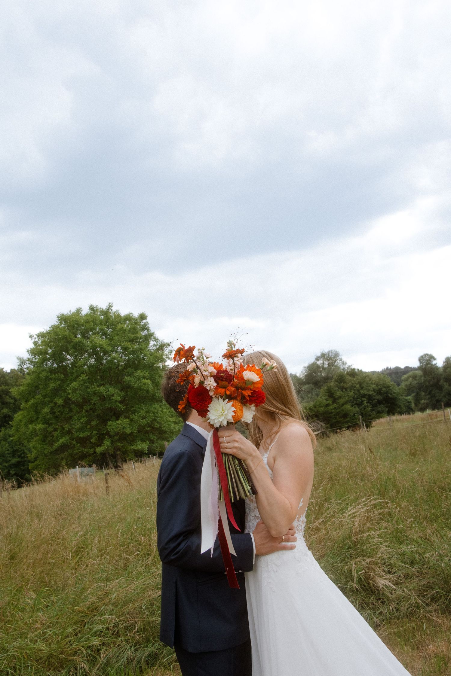 A couple shares a tender moment in a meadow with orange lilies covering their faces on their wedding day.