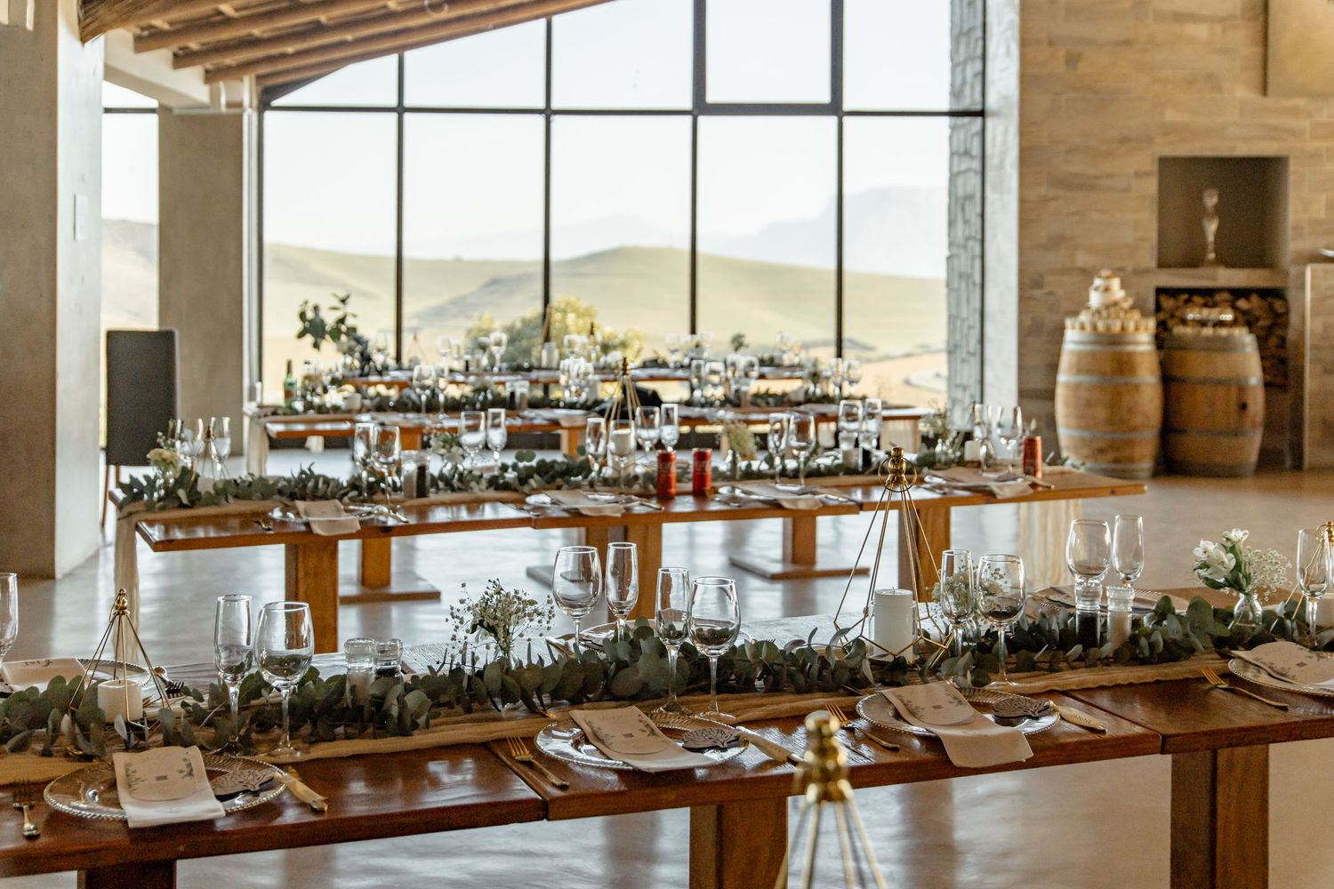 Long rustic wooden tables set for an elegant wedding reception with wine barrel decor and large windows overlooking countryside.