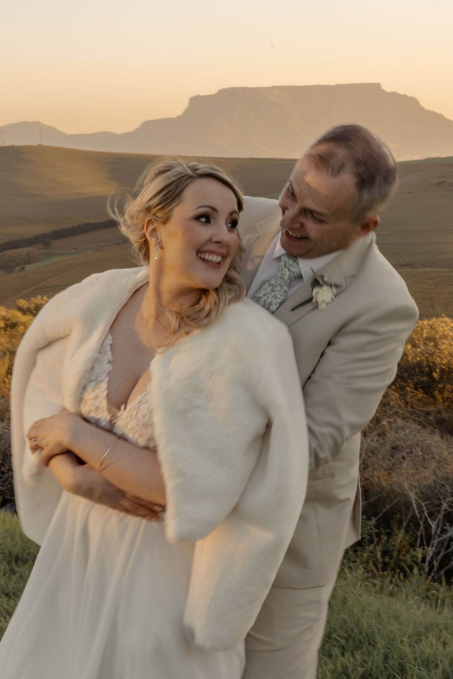 A couple shares a joyful moment during their wedding photoshoot in front of majestic mountain scenery at sunset.