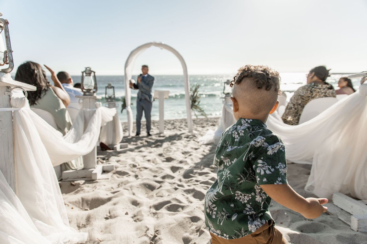 A young child in a tropical shirt looks up during a beach wedding ceremony with flowing white dresses in the background.