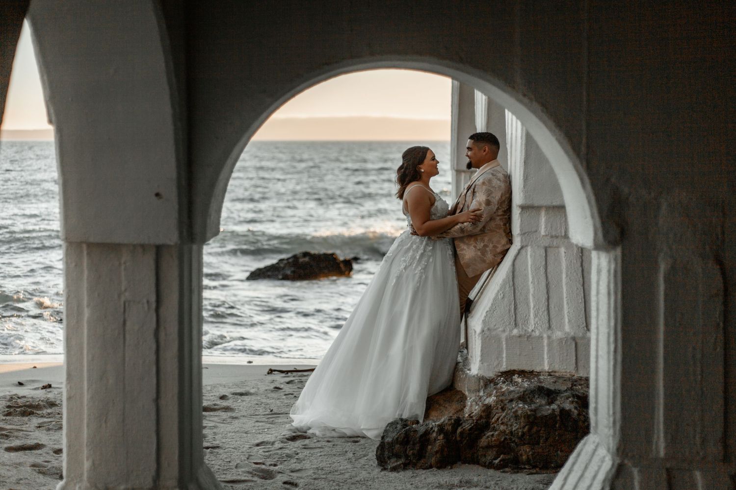 A romantic wedding couple stands in an arched seaside walkway at sunset with ocean waves crashing against rocks.