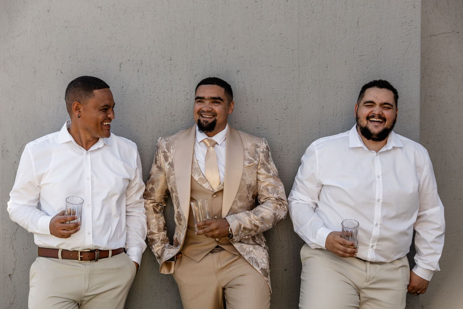 Three stylishly dressed men in white shirts and beige tones laugh together while leaning against a grey wall.