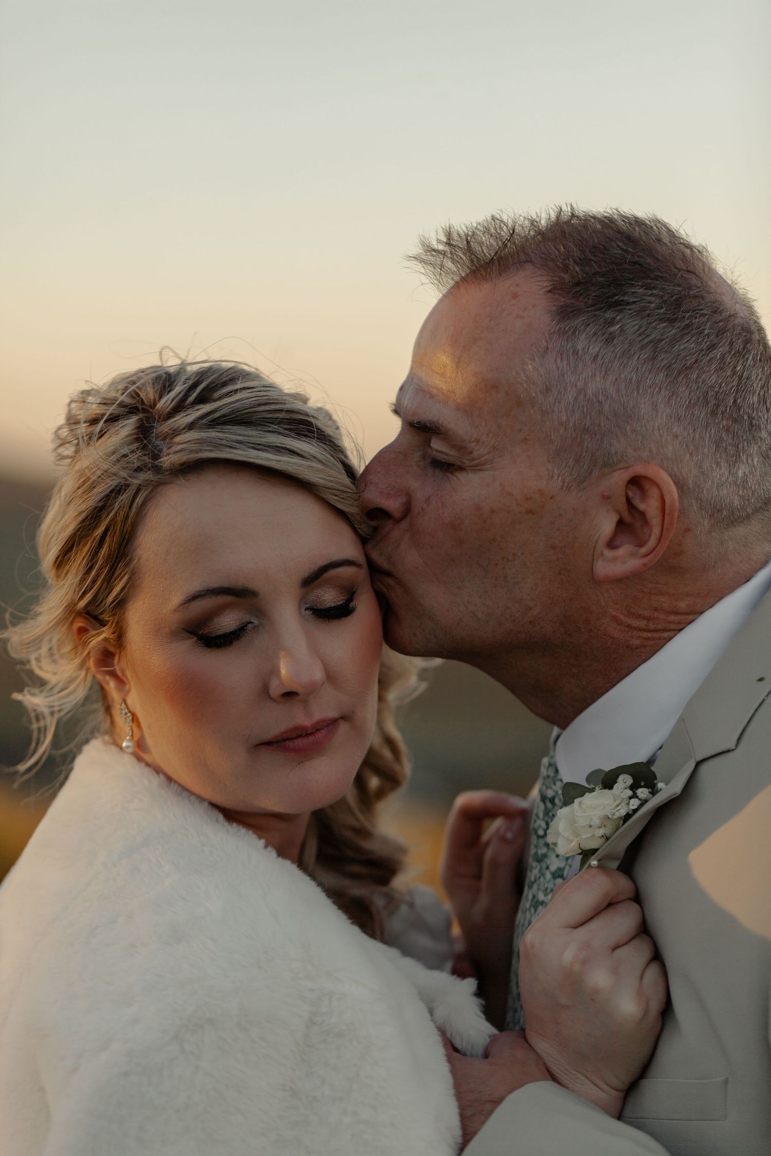 Couple shares intimate moment at sunset with groom kissing bride's forehead while she holds his boutonniere.