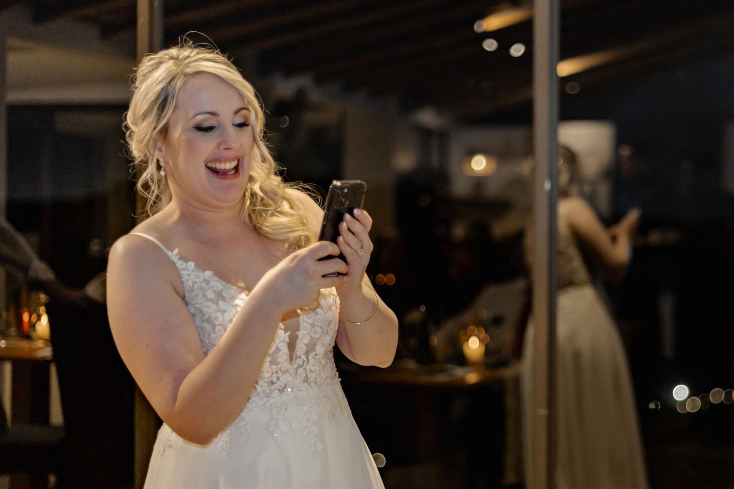 A bride in a white lace wedding dress laughs while looking at her phone during an evening reception.