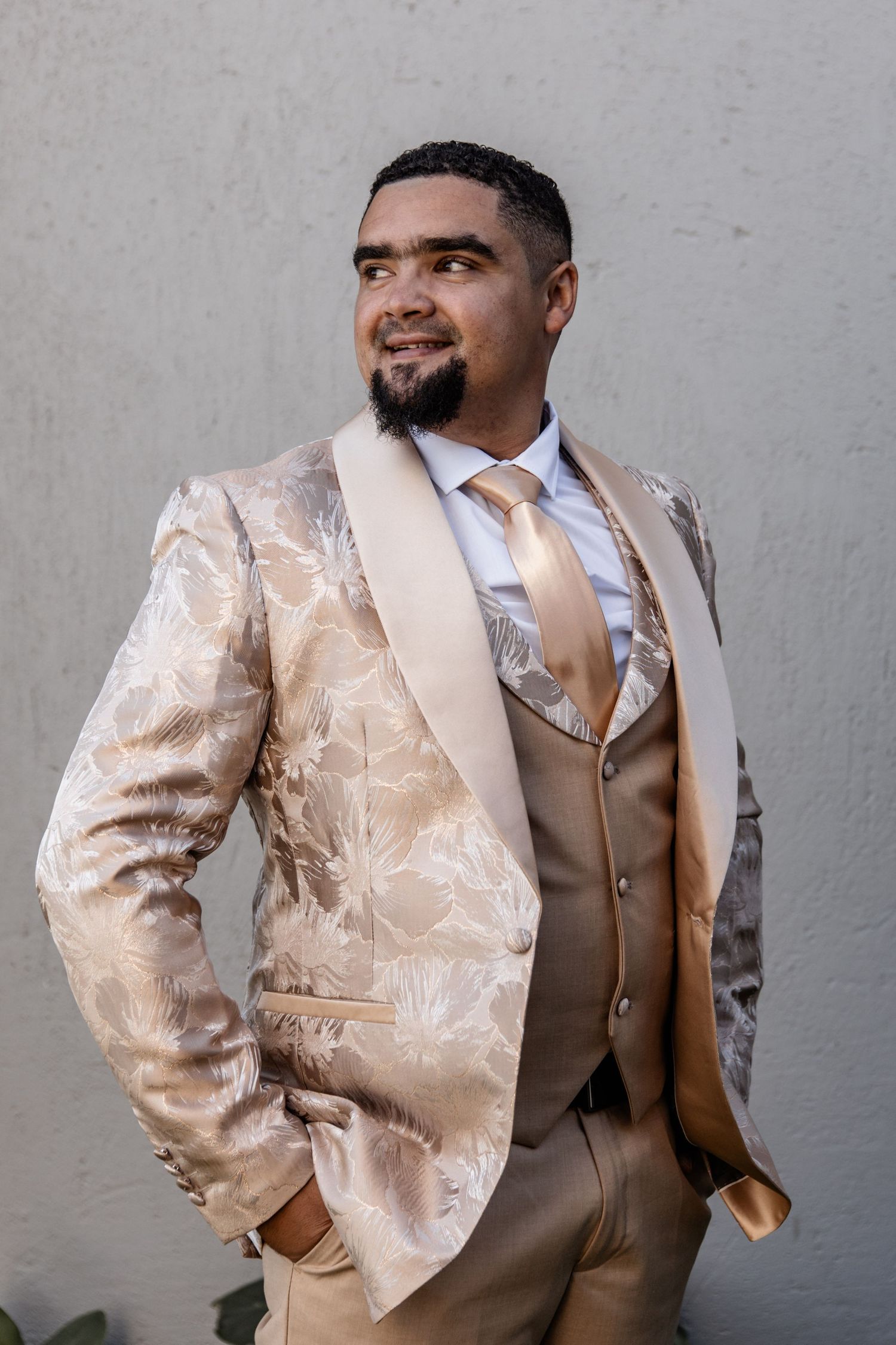 A person wearing an elegant cream floral patterned blazer with tan vest and peach tie stands confidently against a white wall.