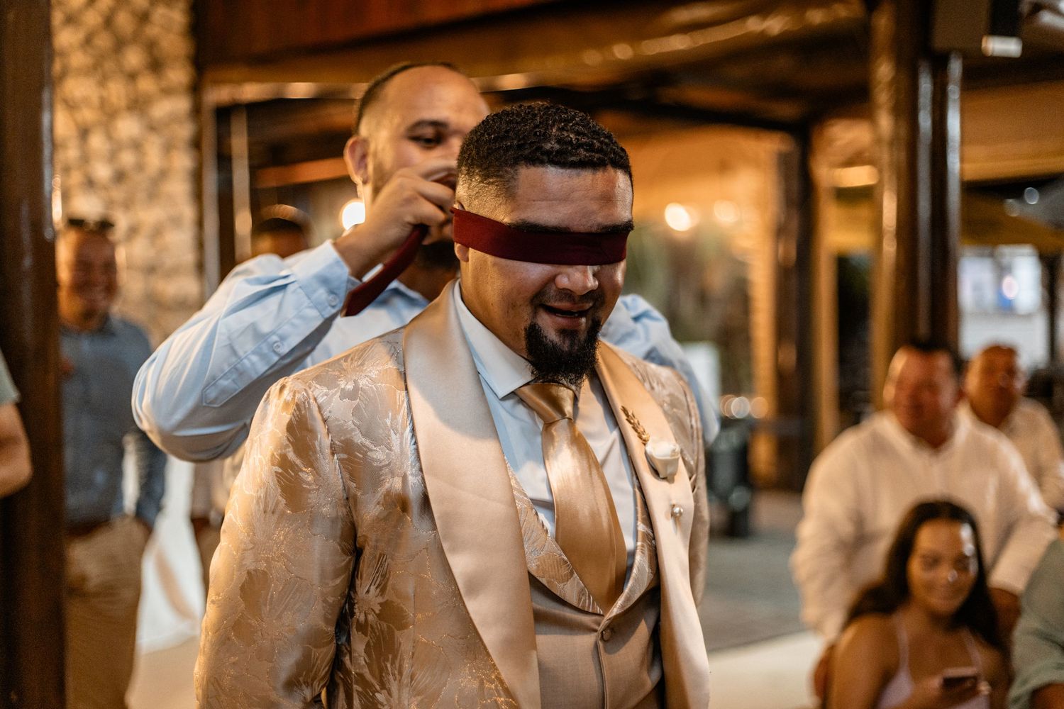 A person in a beige suit has a blindfold put on while standing in an elegant indoor venue with warm lighting.