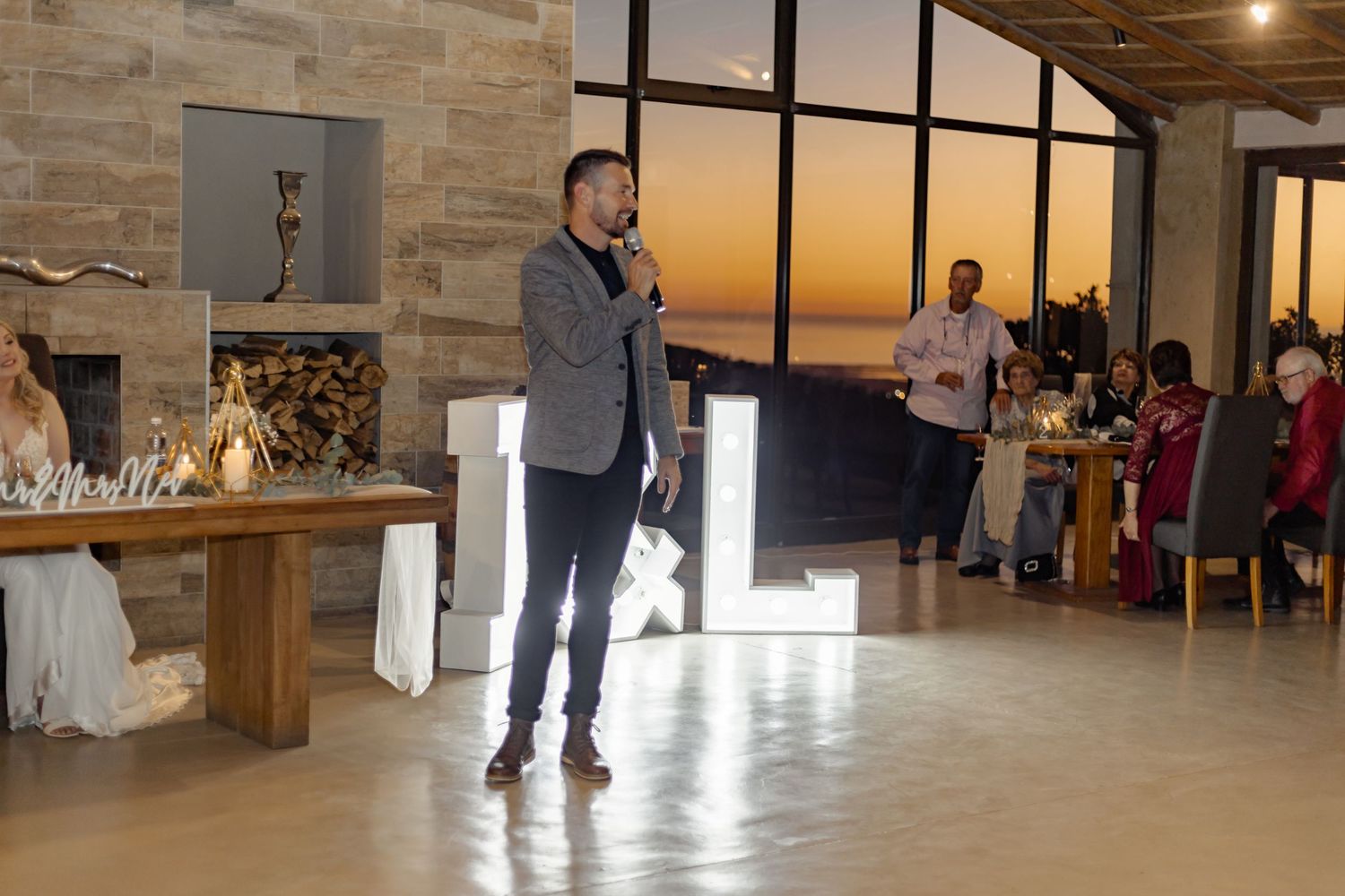 A speaker stands in an open event space with stone walls and wooden tables during a gathering at sunset.