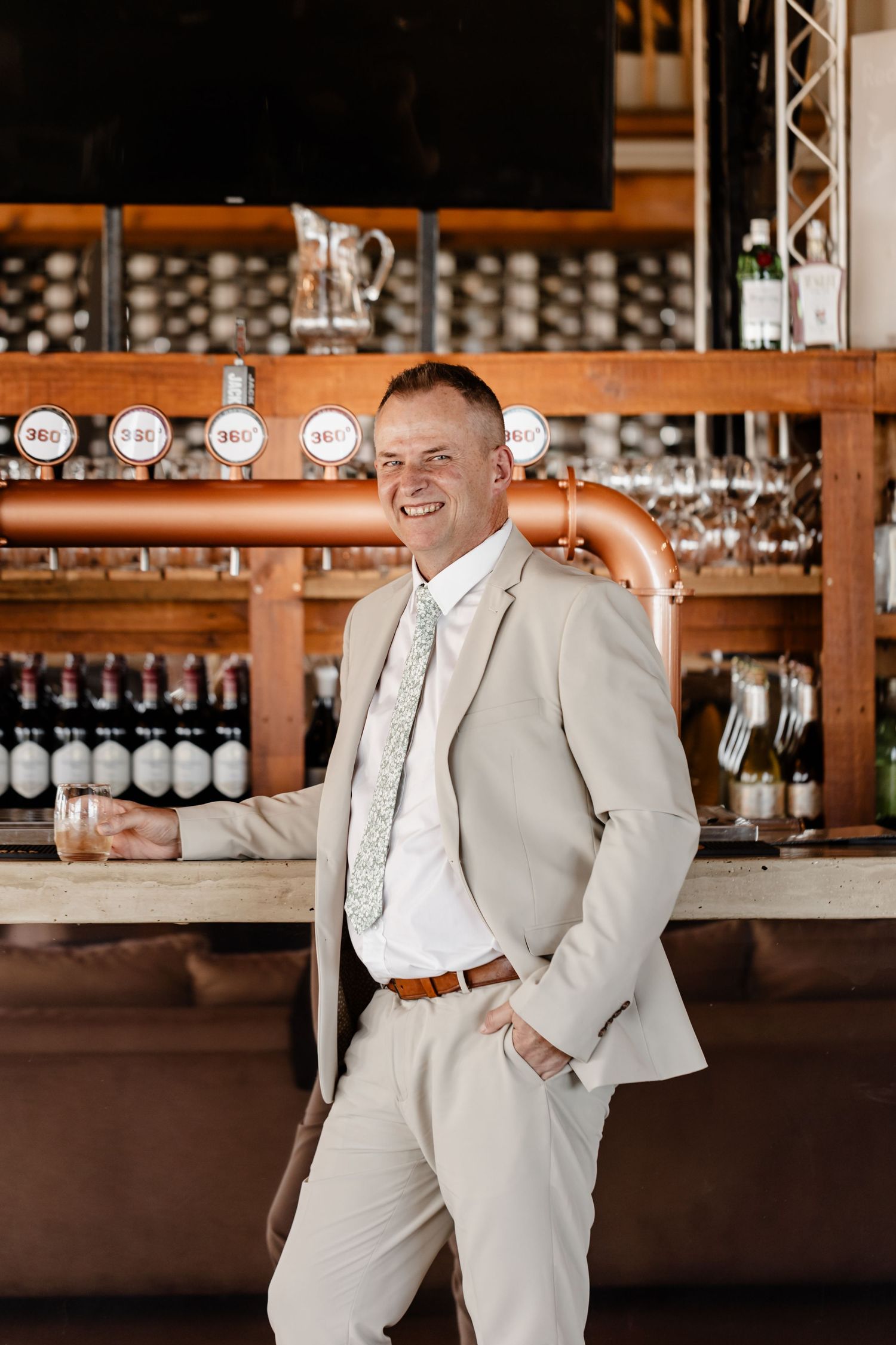 A person wearing a light beige suit and tie leans casually against a bar with wine bottles and glassware displayed behind them.