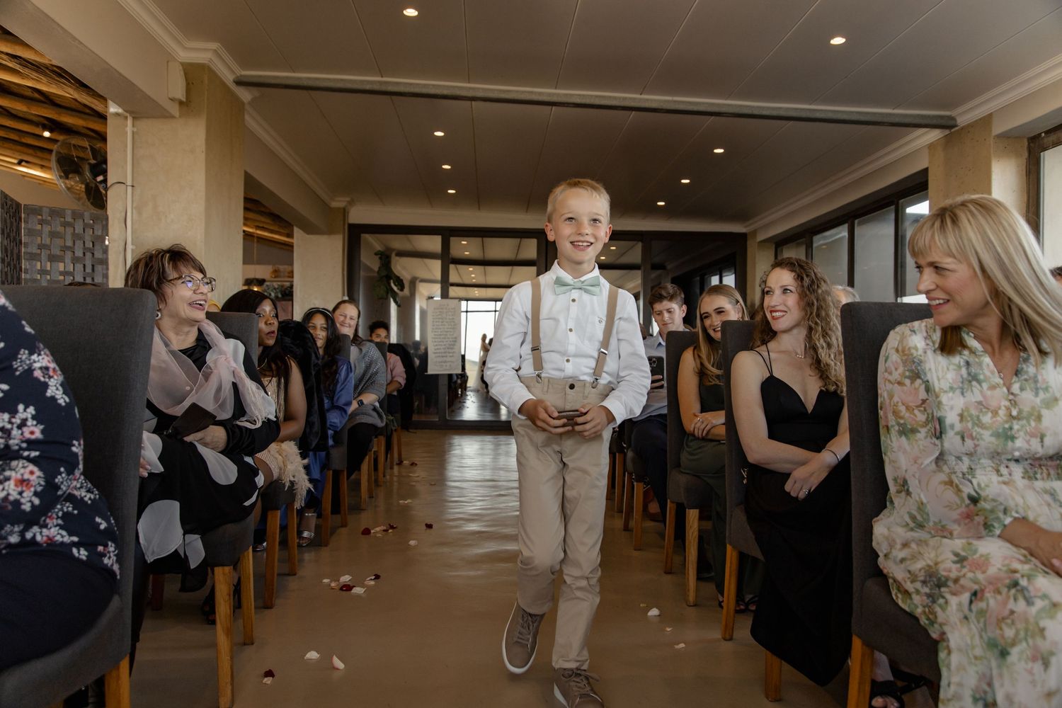 A young child in suspenders walks down an aisle at an indoor event while guests in formal attire watch from both sides.