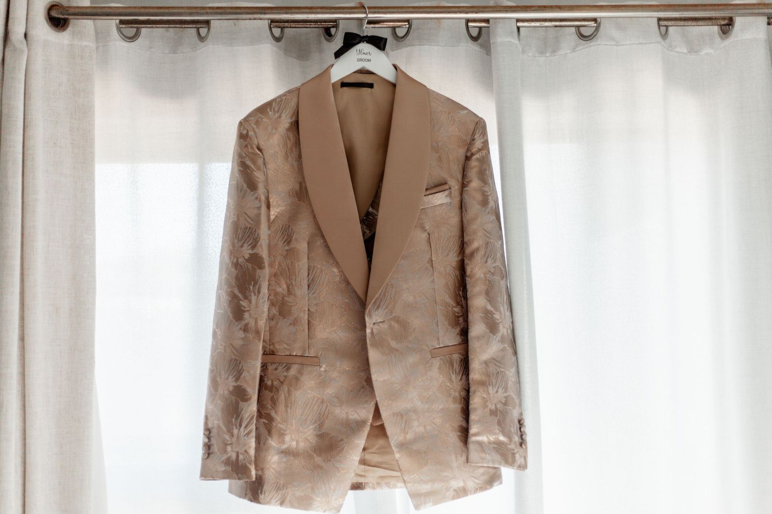 Elegant beige velvet blazer hanging on curtain rod against white sheer curtains in natural light.