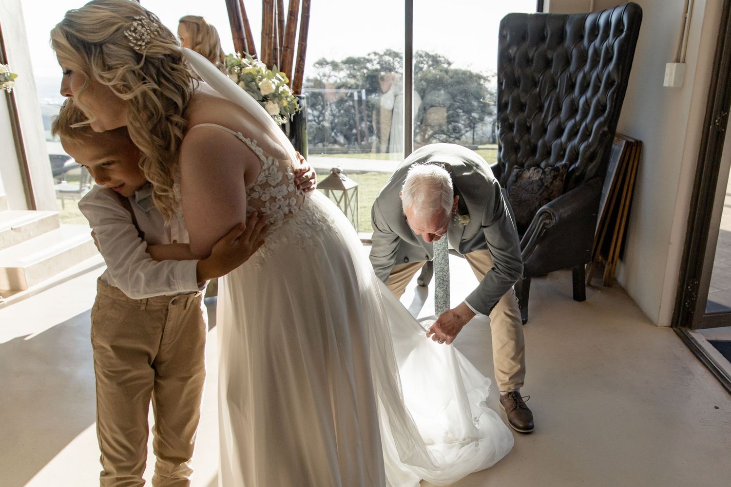Three people in a bright sunlit room with one in a gown getting help adjusting their dress.