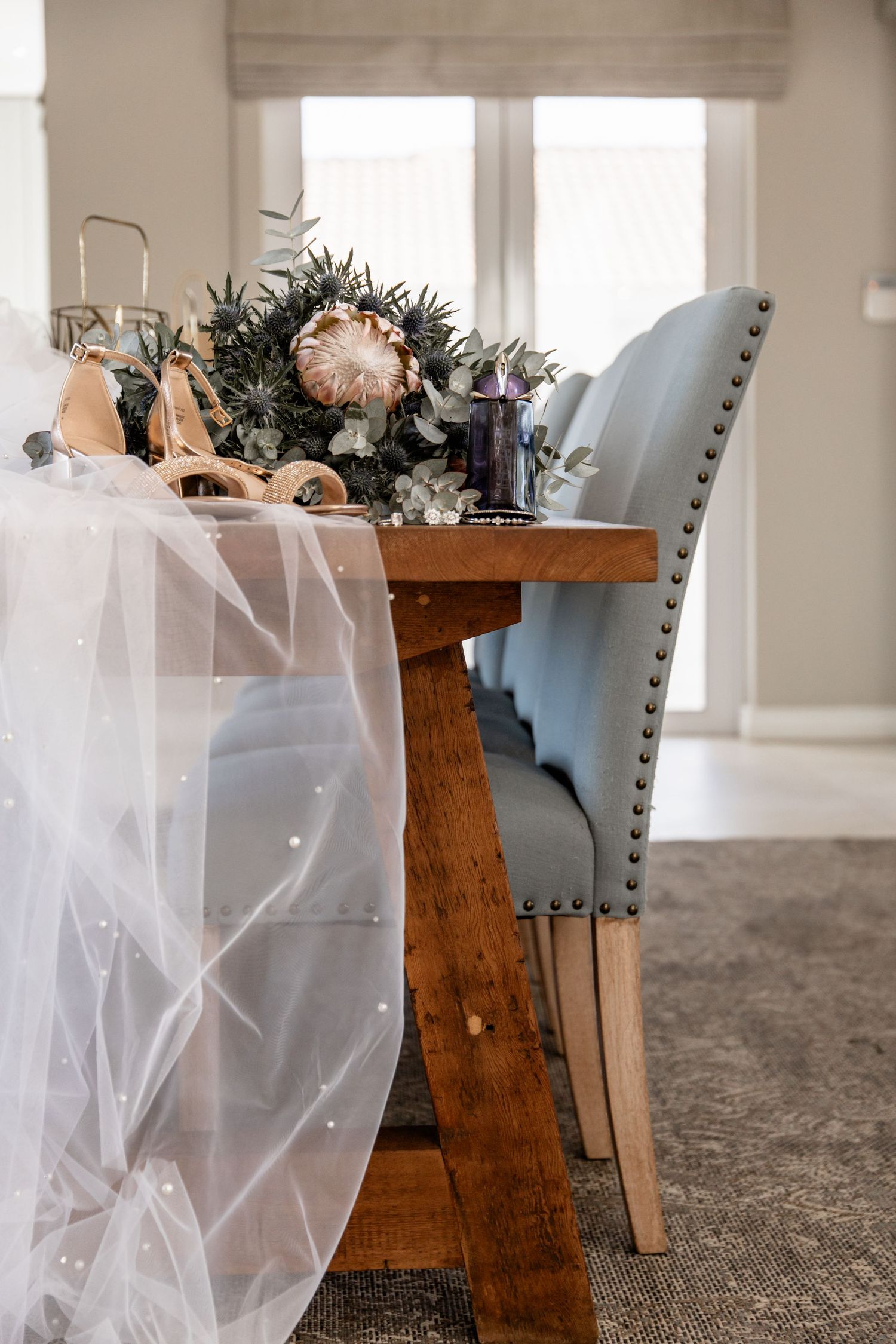 Elegant table setting with delicate tulle draping, rustic wood table, blue upholstered chair and floral centerpiece arrangement.
