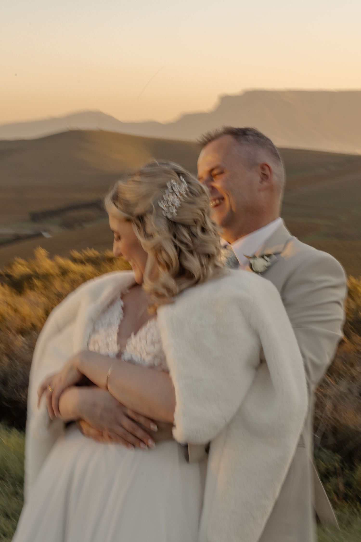 A couple embraces during golden hour on a mountainside, with the bride wearing a white gown and fur shawl at their sunset wedding.