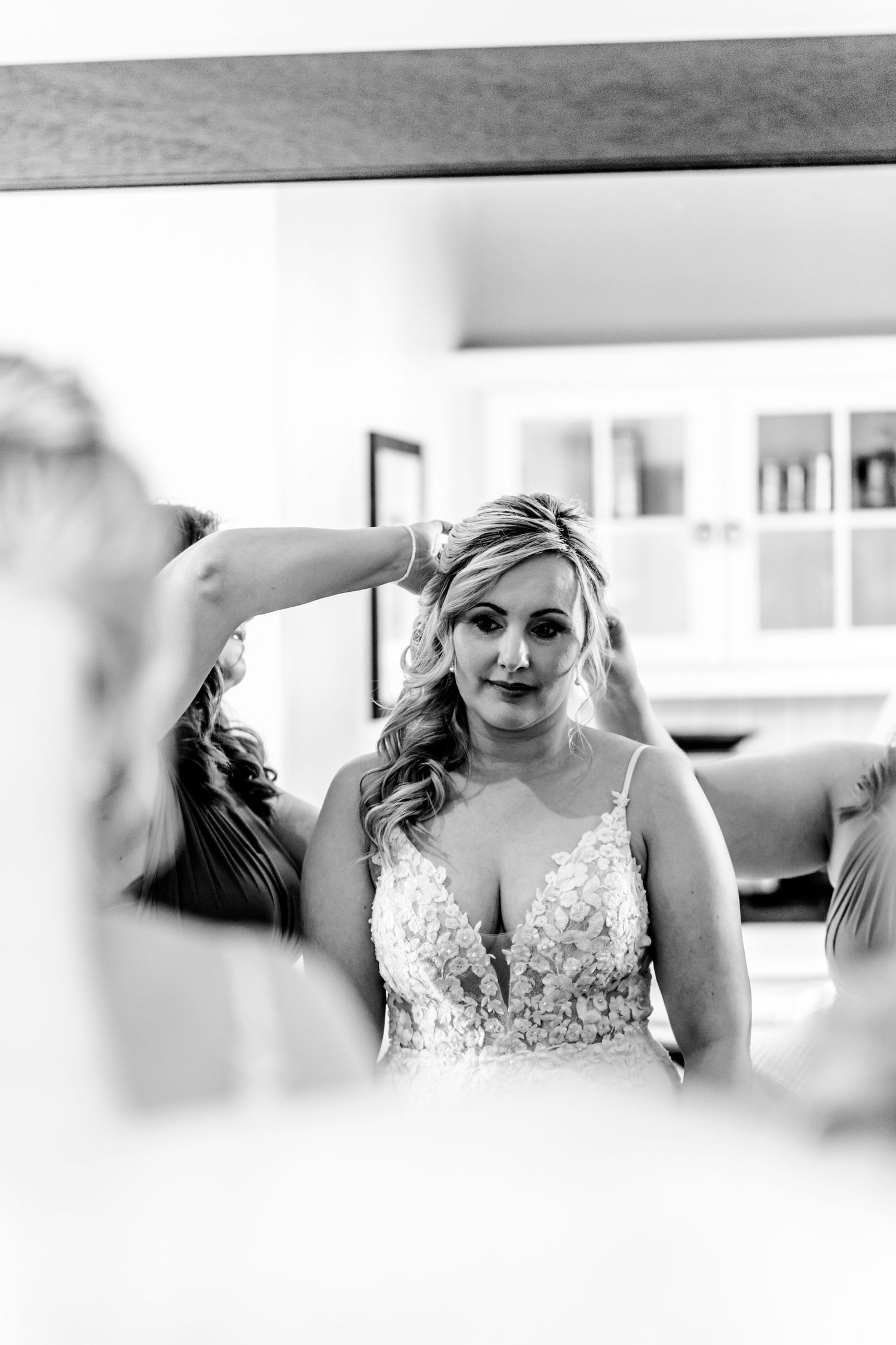 Black and white mirror reflection showing woman in lace detailed wedding gown with wavy blonde hair being styled.