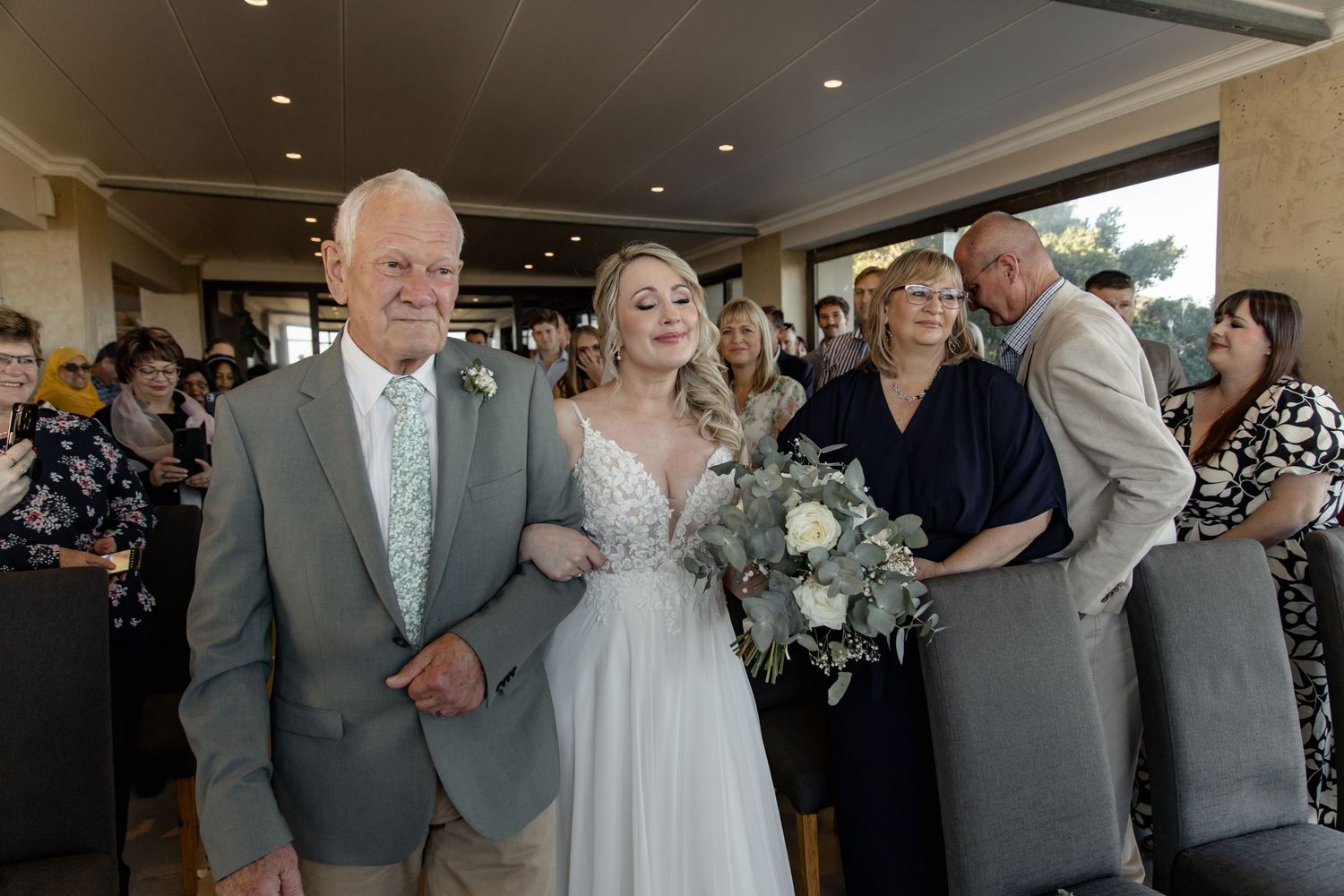 A couple smiles while surrounded by wedding guests in an elegant indoor venue with warm lighting.