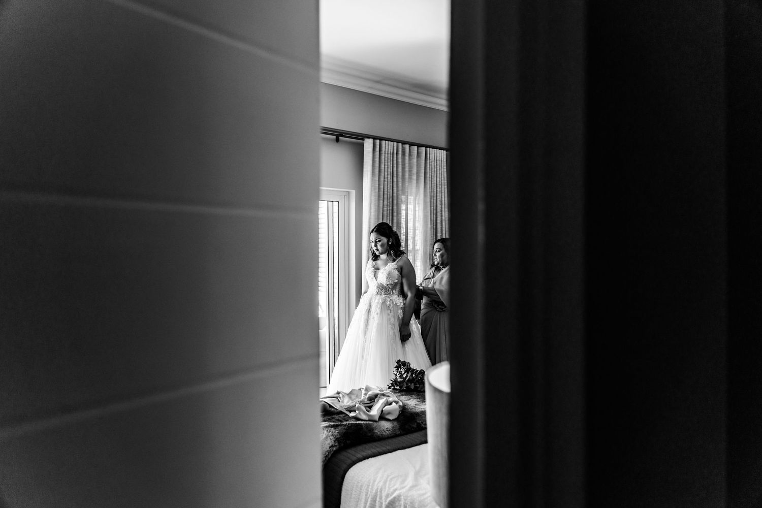 Black and white view through doorway showing silhouetted figure in wedding dress getting ready in sunlit hotel room.