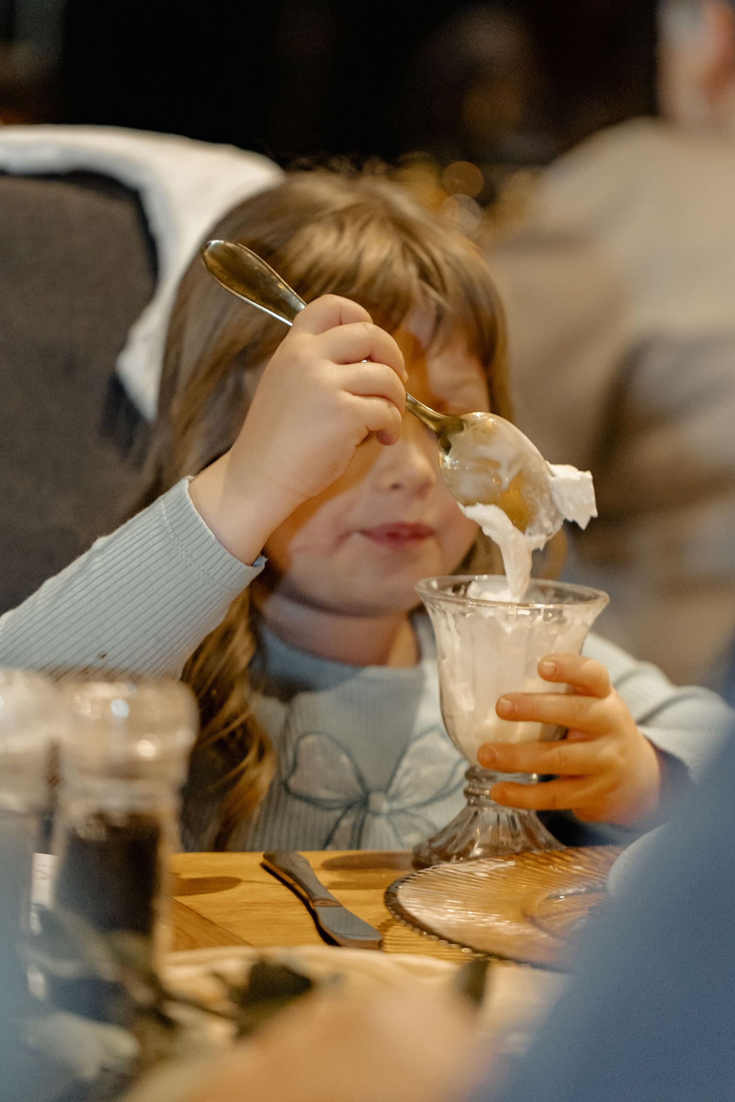 Child wearing light blue sweater enjoying ice cream dessert at restaurant table with dreamy expression.