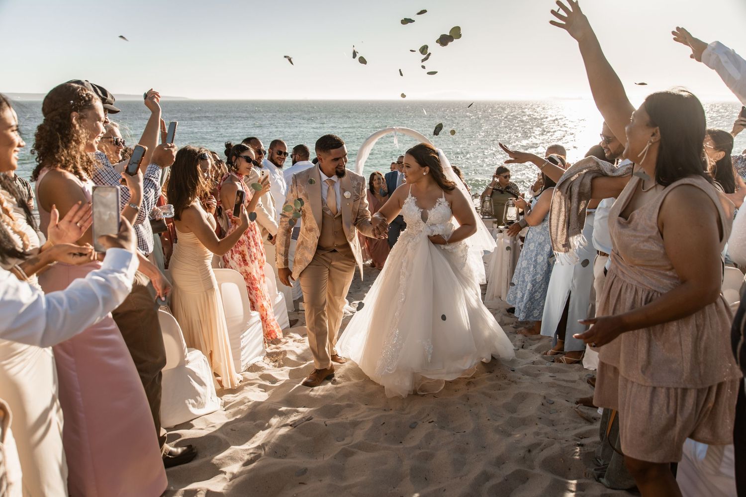 Wedding celebration on a beach with guests throwing petals as newlyweds walk between joyful crowd at sunset.