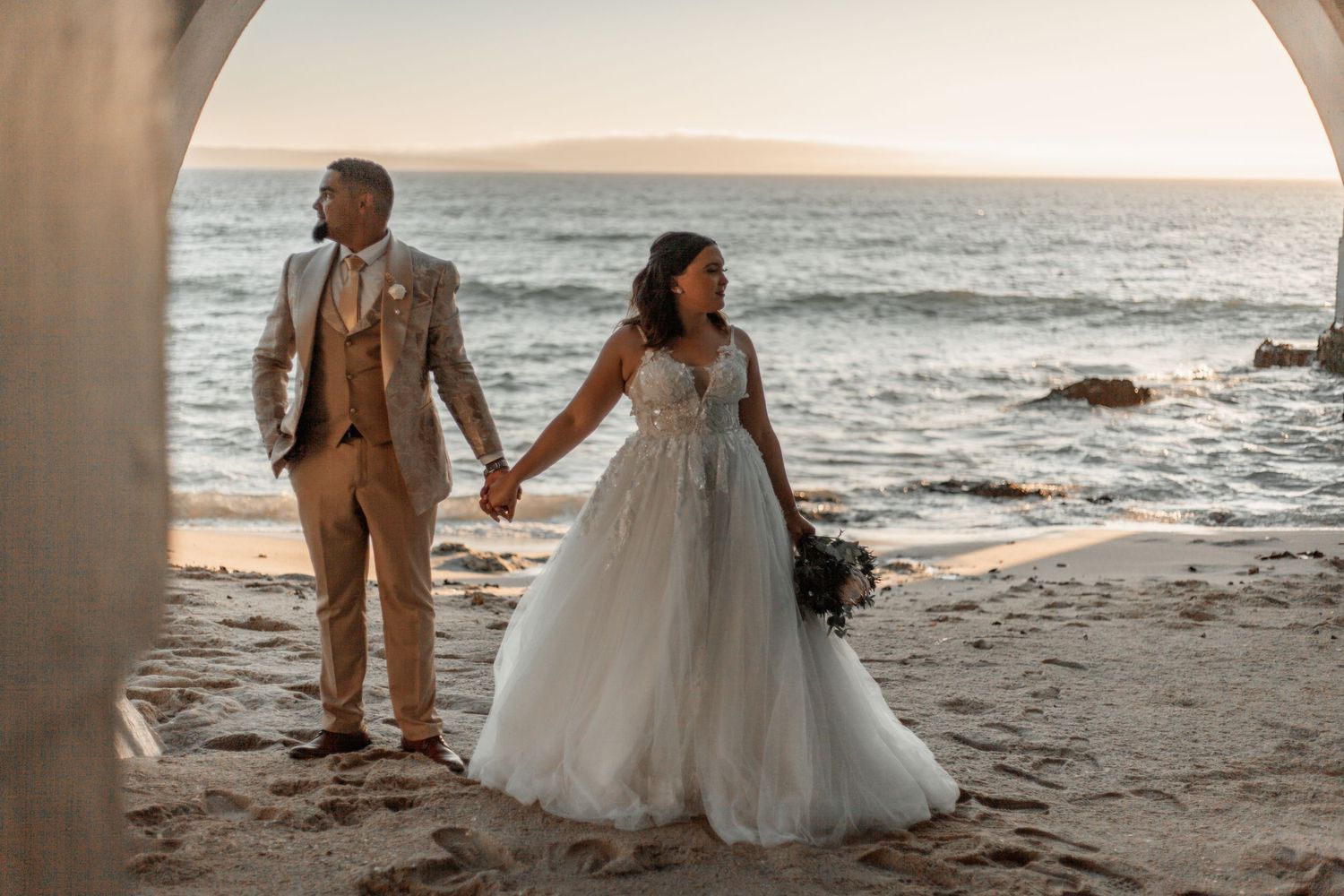 Newlywed couple in formal attire holding hands while walking on sandy beach at sunset with ocean waves in background.