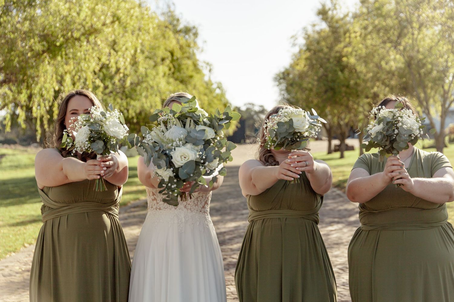 A bridal party in sage green dresses holds bouquets of white and green flowers in front of their faces outdoors.
