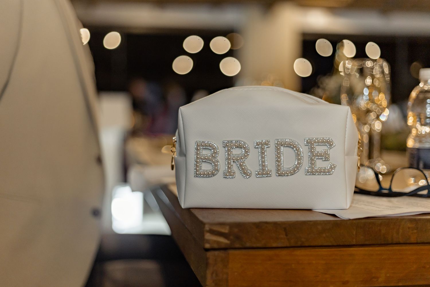 A white wedding sign with 'BRIDE' written in silver glittery letters sits on a wooden table with blurred lights in background.