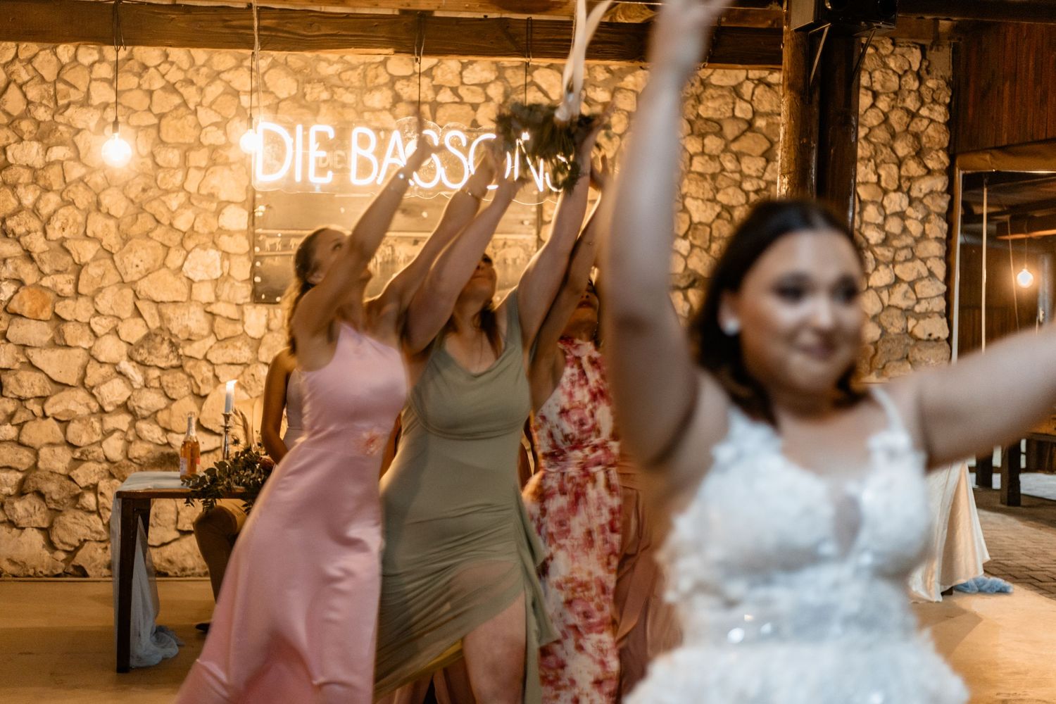 Dancers celebrating at a rustic wedding reception with stone wall backdrop and neon 'Die' signage.