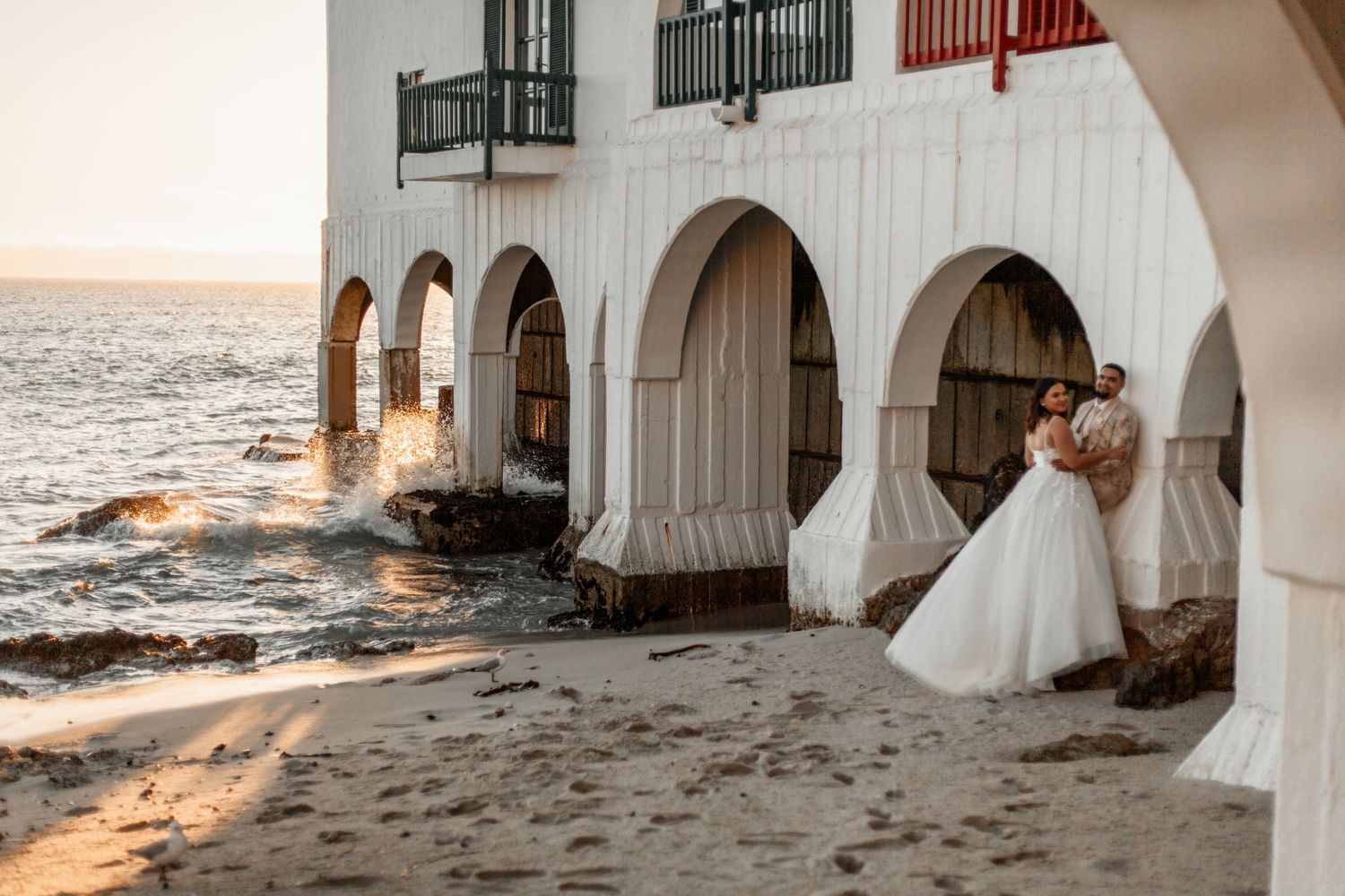 Couple embraces under white Mediterranean arches by the ocean at sunset, waves crashing against shore.