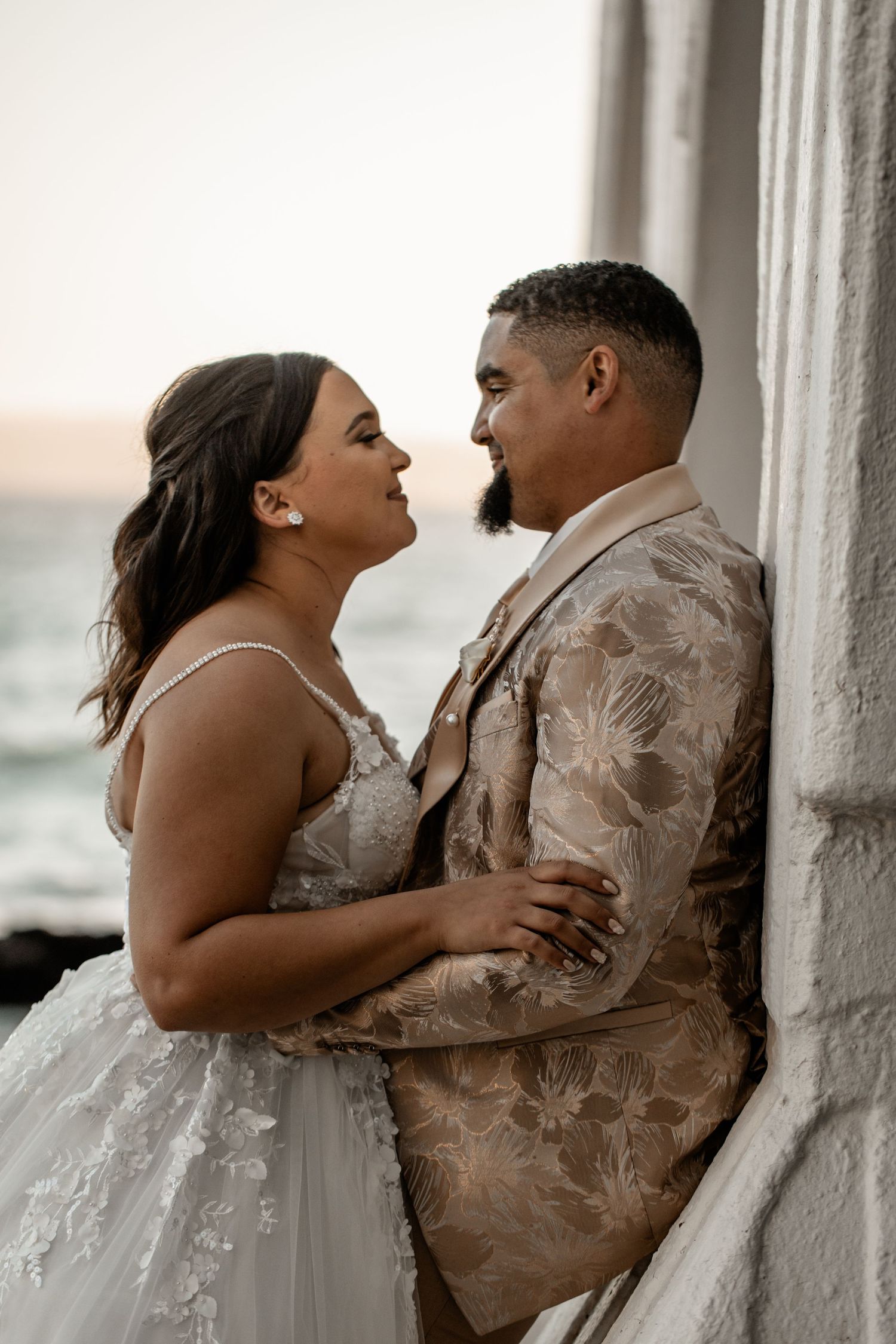 A joyful couple share an intimate moment in their wedding attire against a bright background.