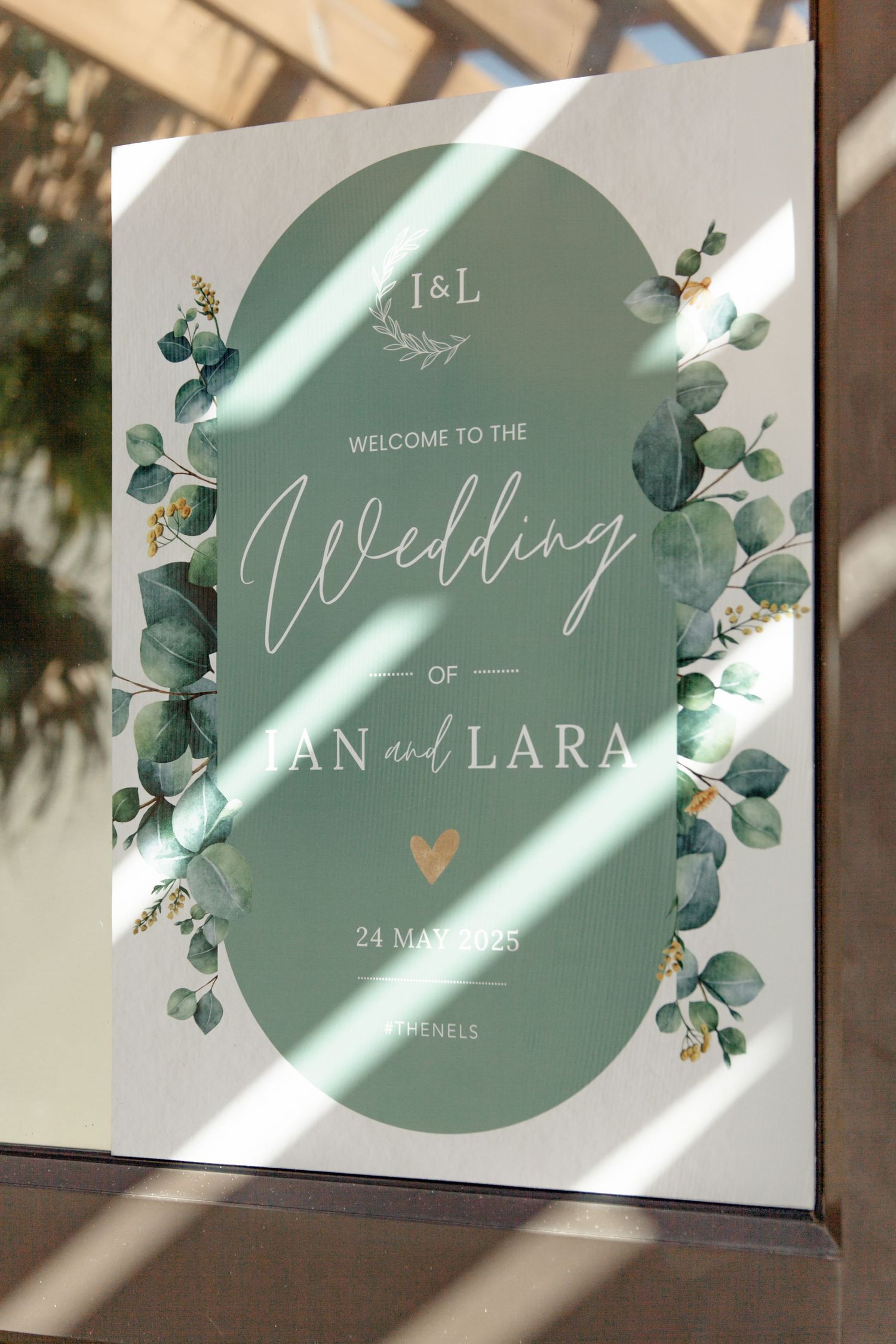 Elegant sage green wedding welcome sign with eucalyptus leaves and gold heart detail shown in natural window light.