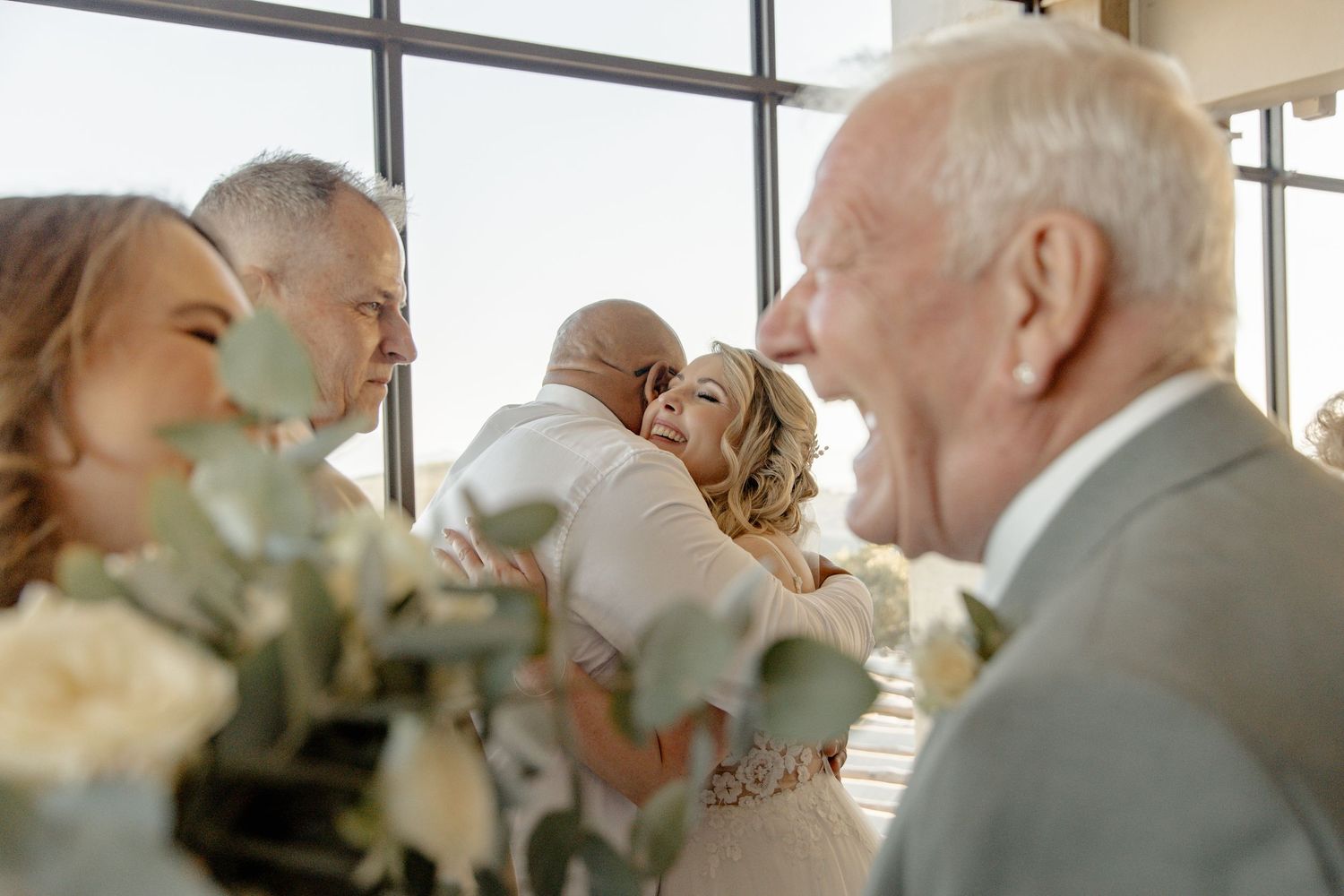 Wedding guests share heartfelt laughter during an intimate indoor reception with soft window lighting and white floral decor.
