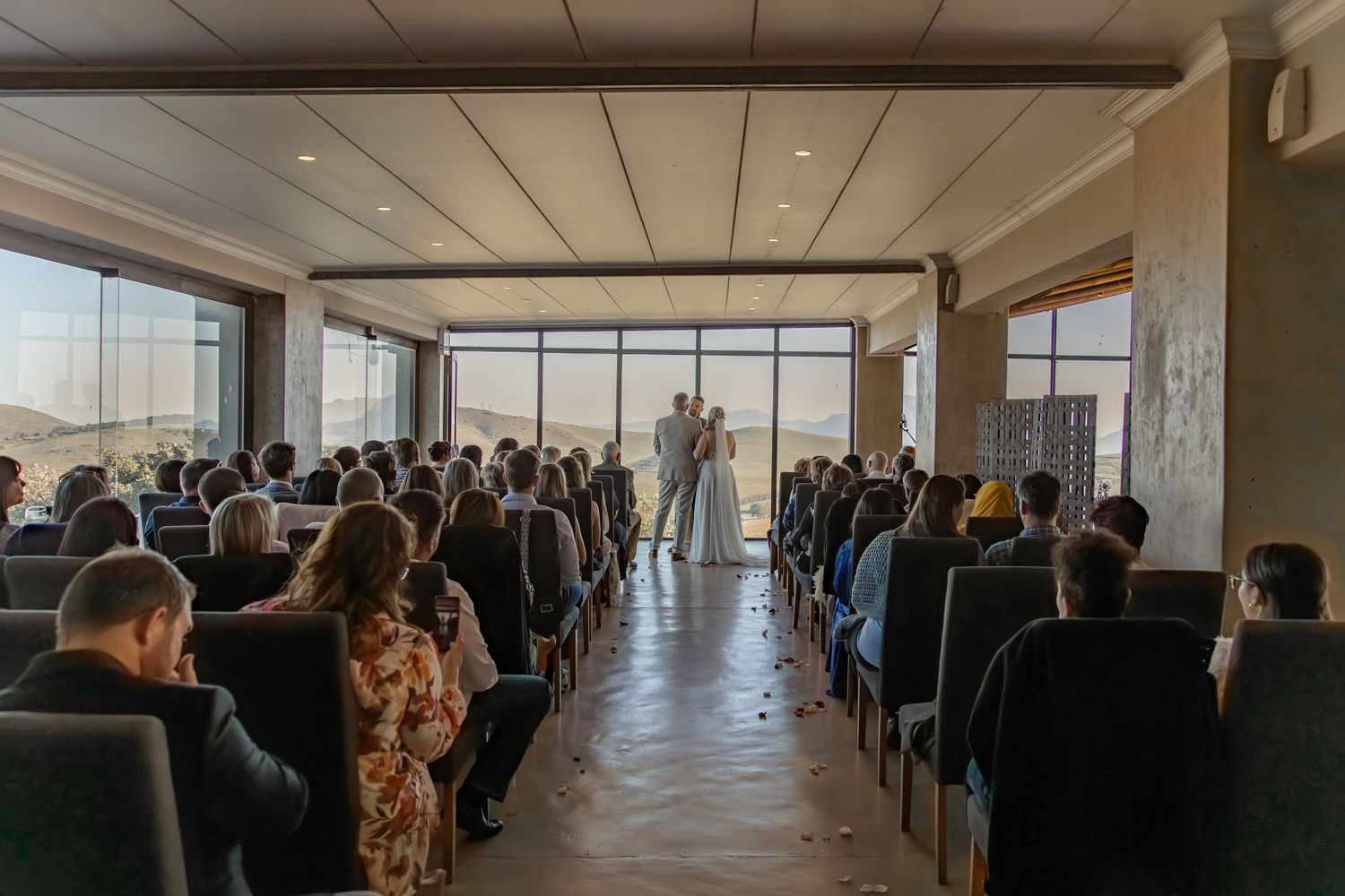 A wedding ceremony taking place in a modern venue with floor-to-ceiling windows and rows of seated guests viewing the ceremony.