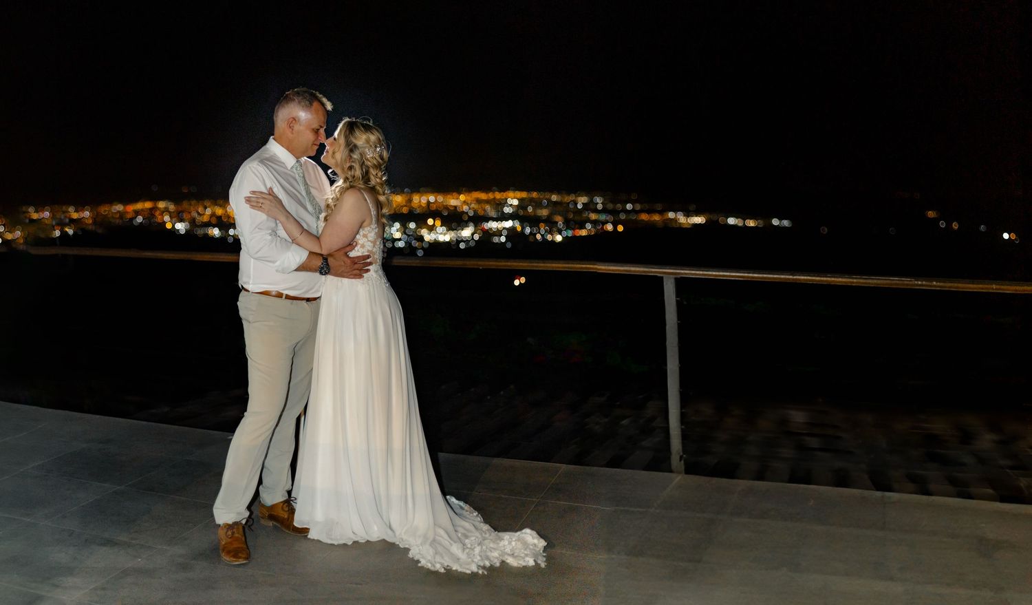 A couple shares an intimate moment on a balcony at night with city lights glowing in the background.