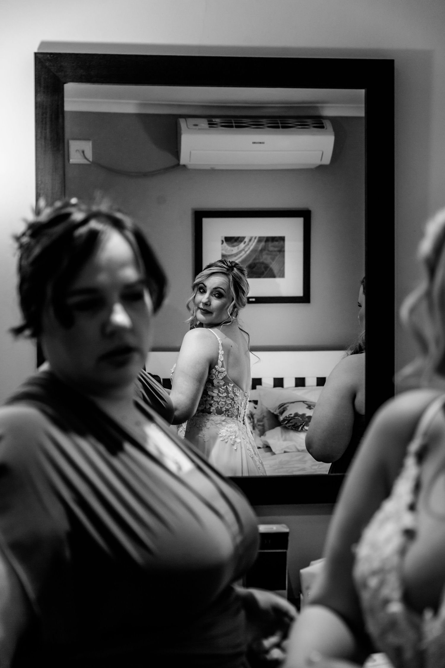 Black and white candid photo showing people getting ready in a room with a mirror and air conditioning unit visible.