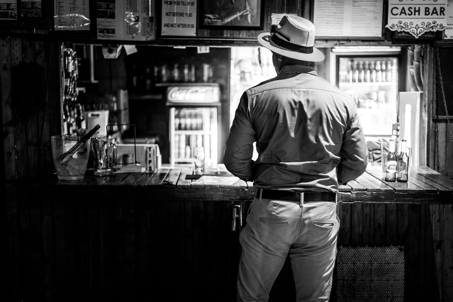 A person in a hat leans against a vintage bar counter in black and white photography.