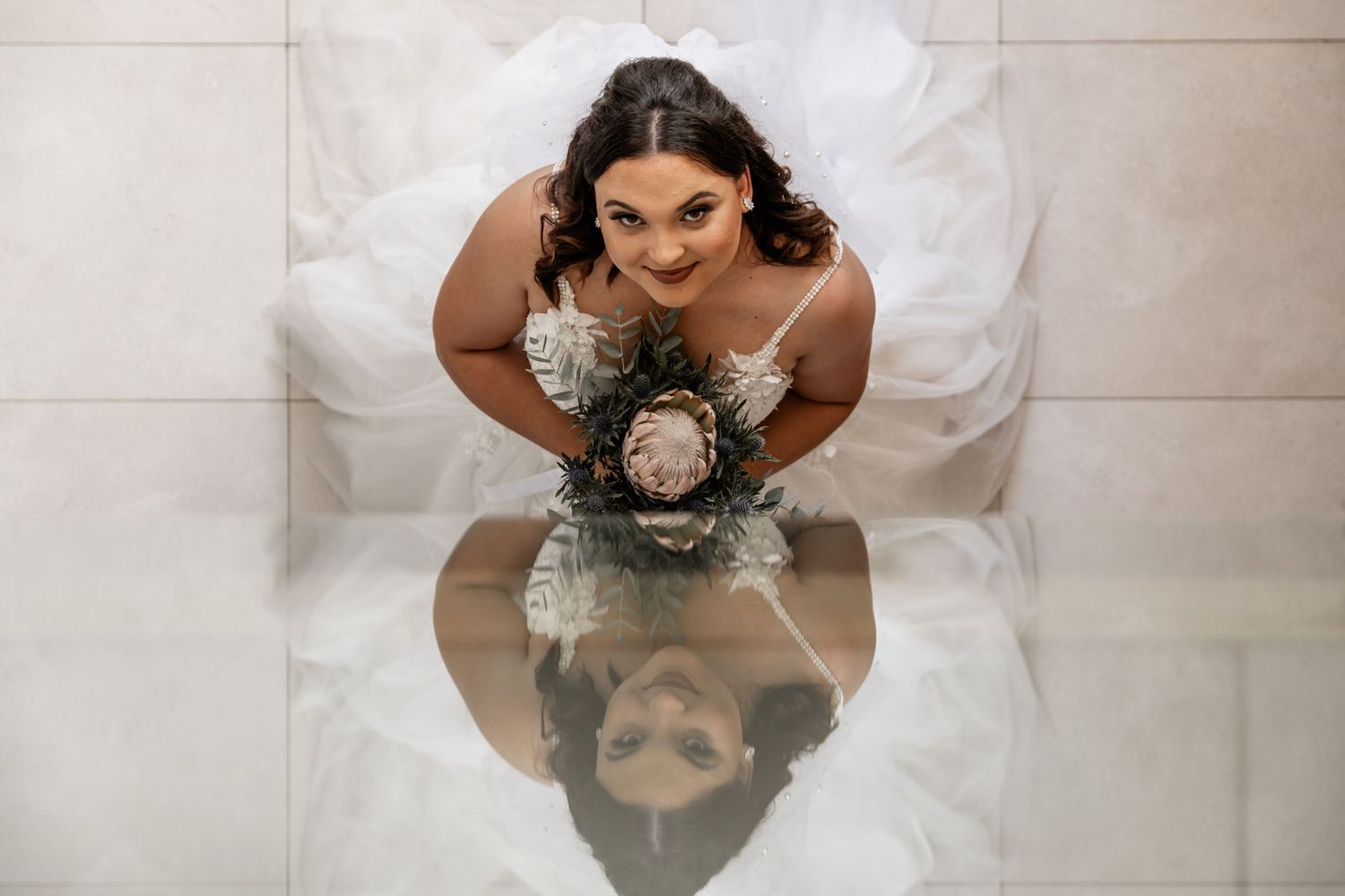 Aerial view of person in white dress looking down at their reflection on polished floor while holding bouquet.