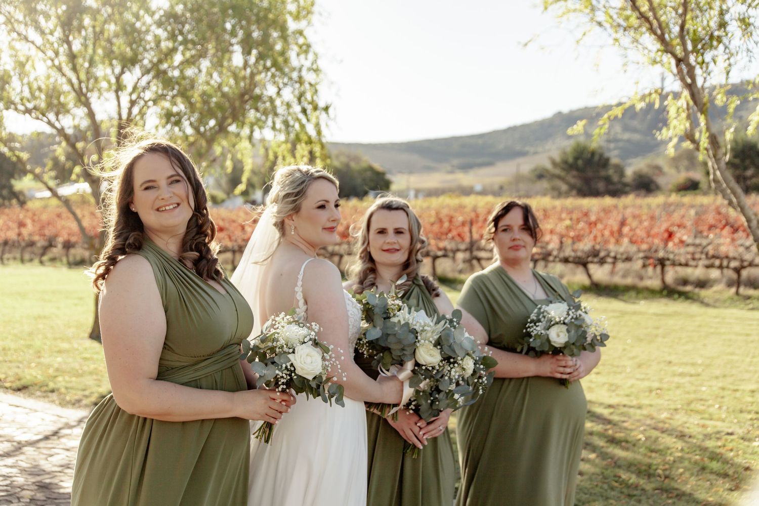 A group stands together on a lawn in front of autumn vineyards wearing elegant sage green and white dresses for a wedding.