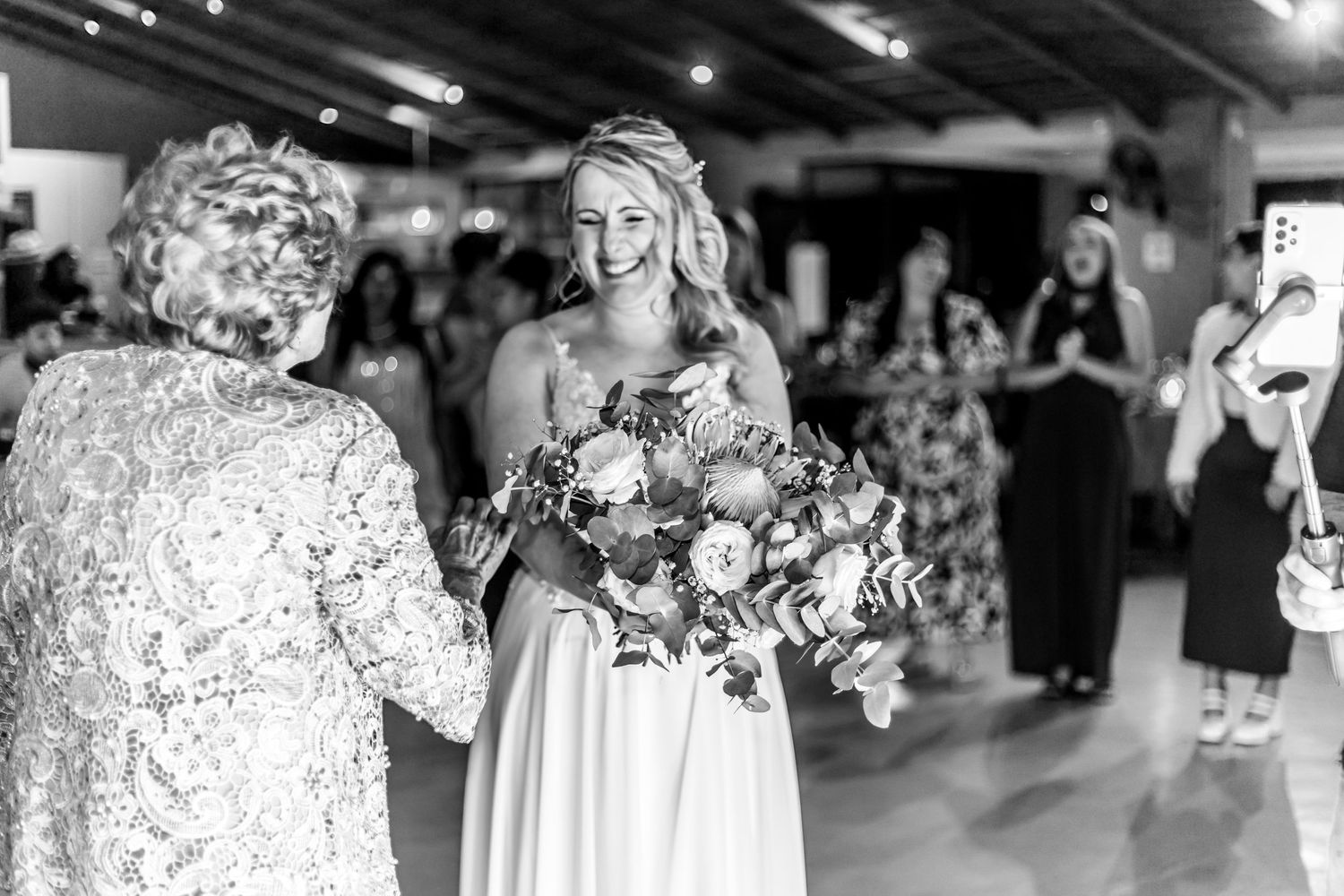 Black and white photo of a bride in a white gown holding a bouquet while engaging with wedding guests at indoor reception.