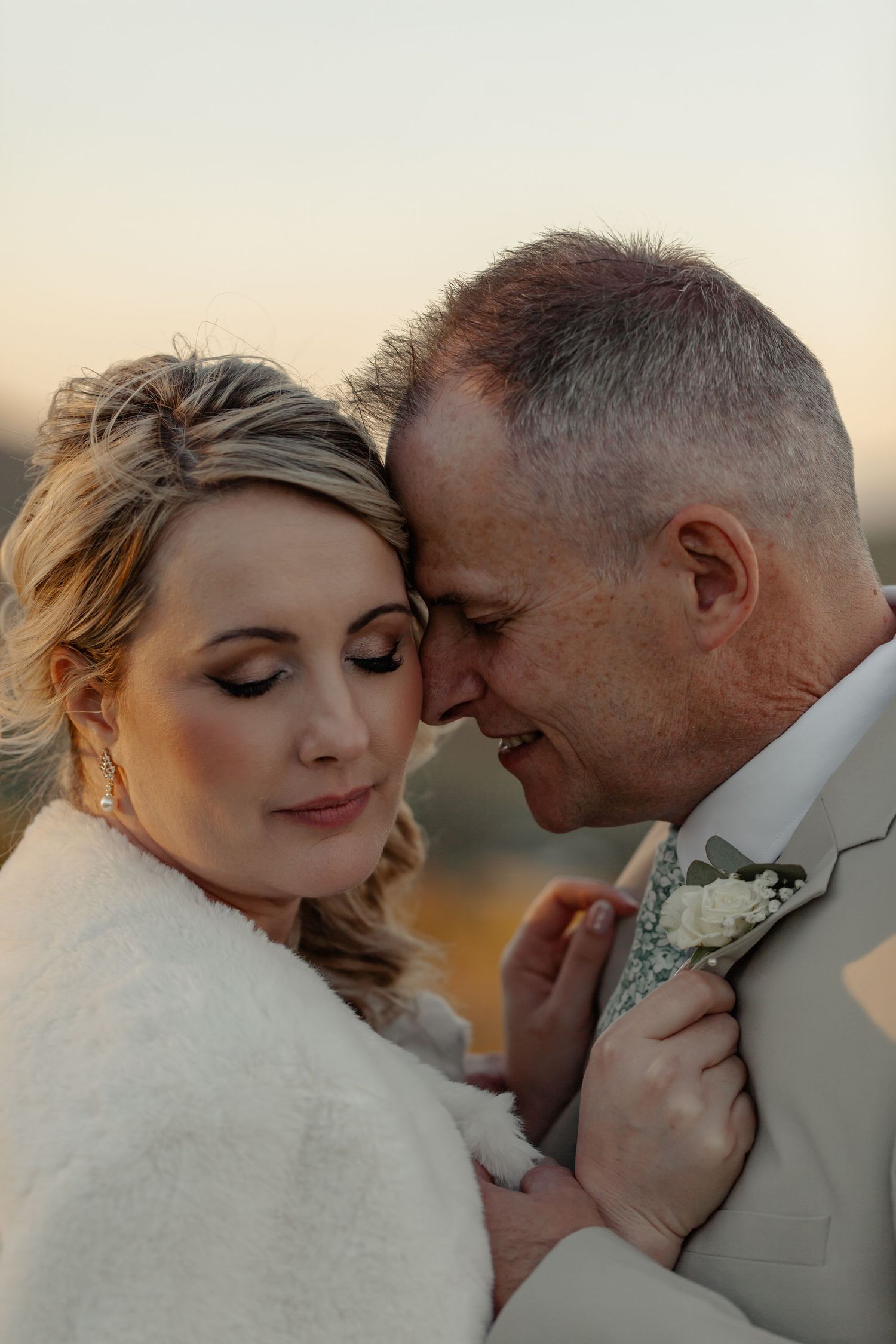 A romantic wedding couple sharing an intimate moment at sunset, with the bride wearing a white shawl and boutonniere.