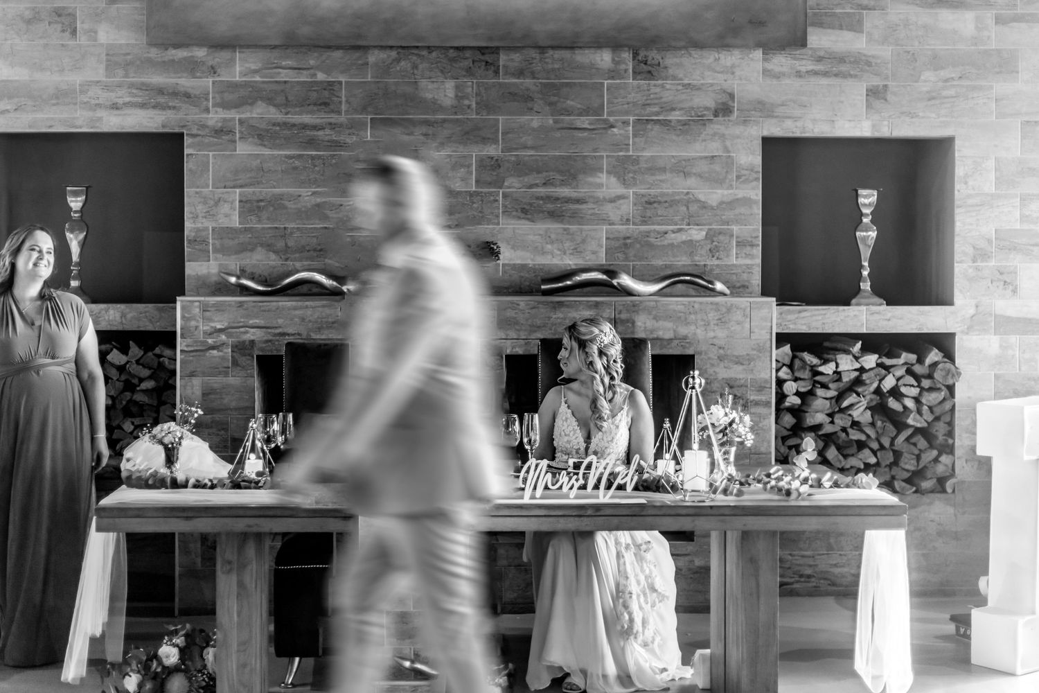 Black and white photo showing a blurred figure walking past a table setup with glassware and decorations against a brick wall.