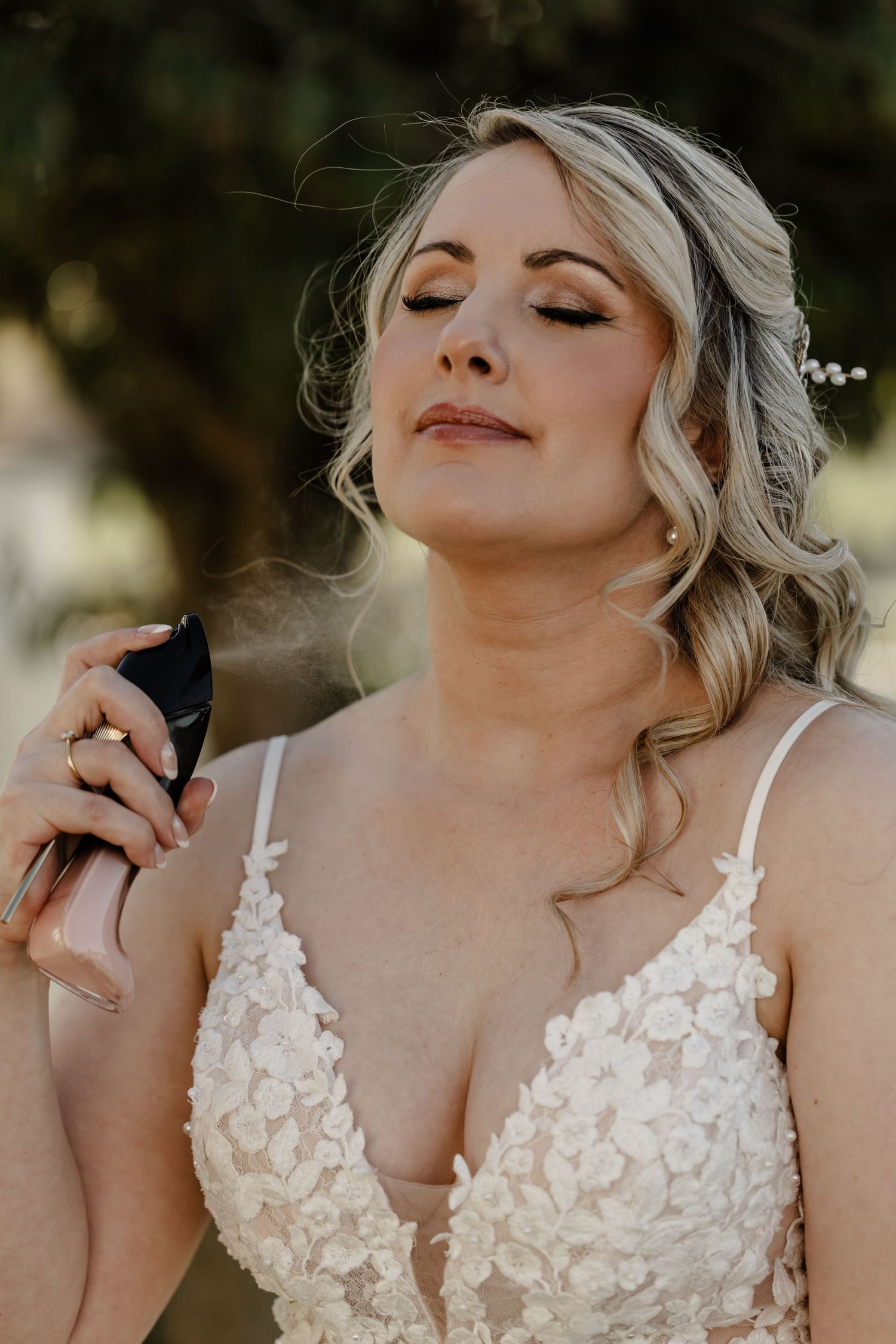 A bride in a white lace wedding dress with blonde curled hair sprays perfume while looking serene with eyes closed.