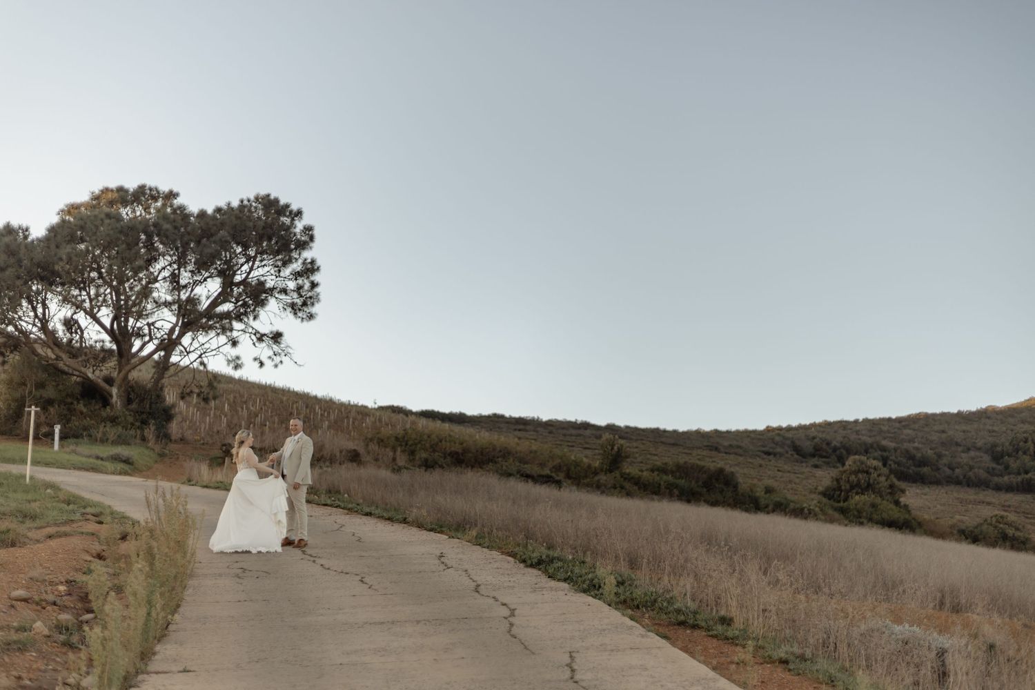 Two people in formal attire walk together down a winding path through rolling hills at sunset.
