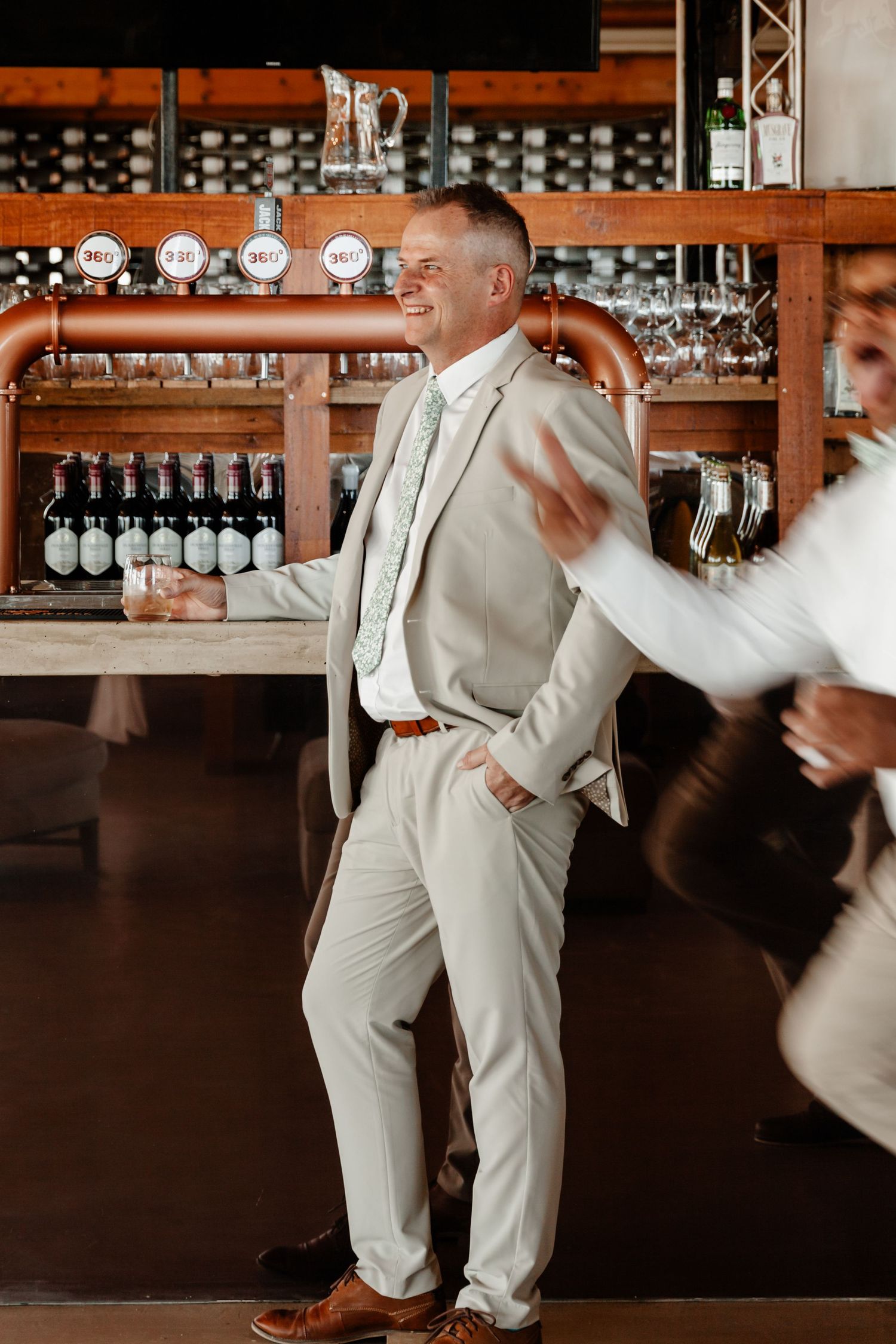 Stylishly dressed person in light beige suit dancing and celebrating at a bar with bottles and glassware displayed.