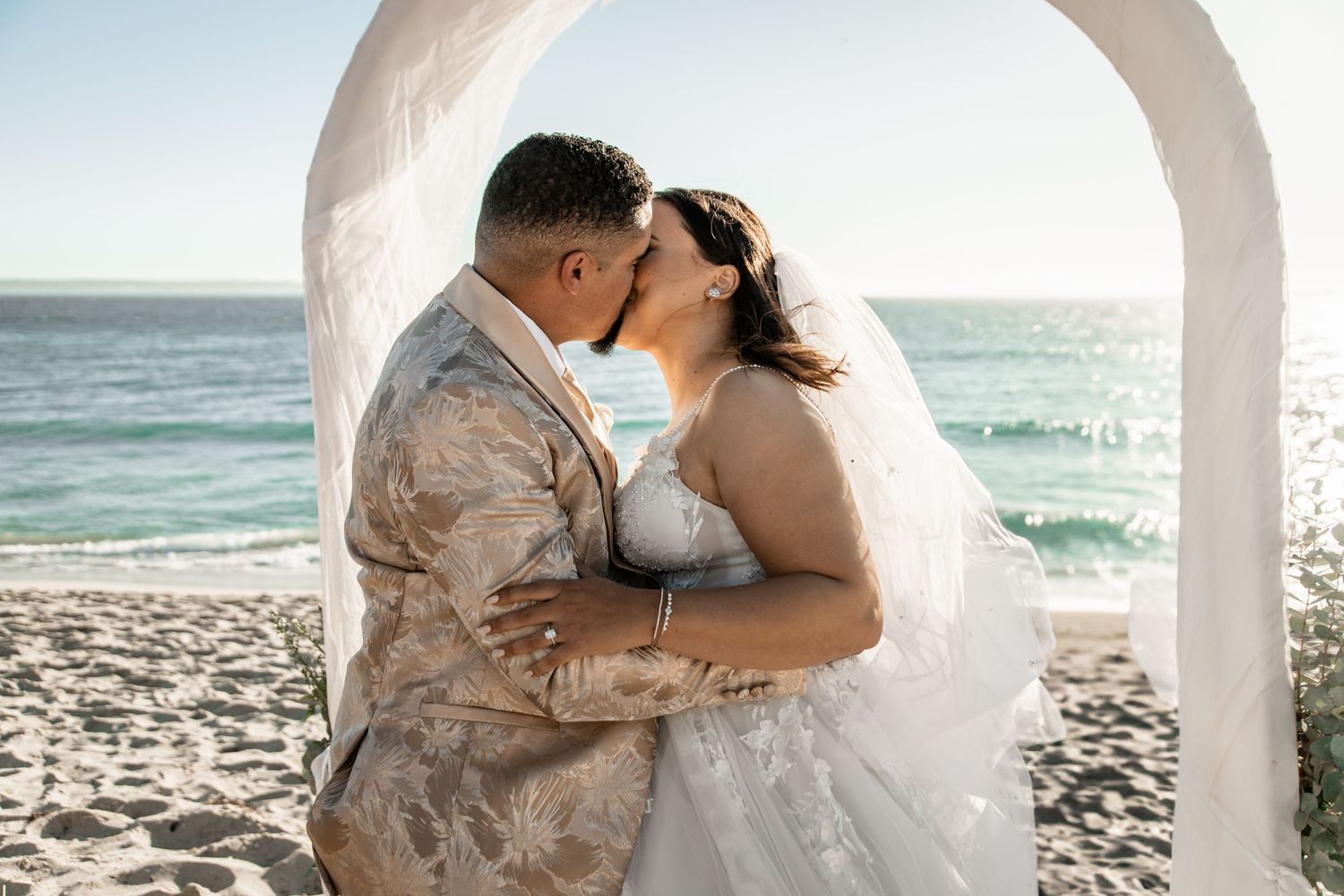 A couple shares a romantic kiss under a white arch on a sunny beach with ocean waves in the background.