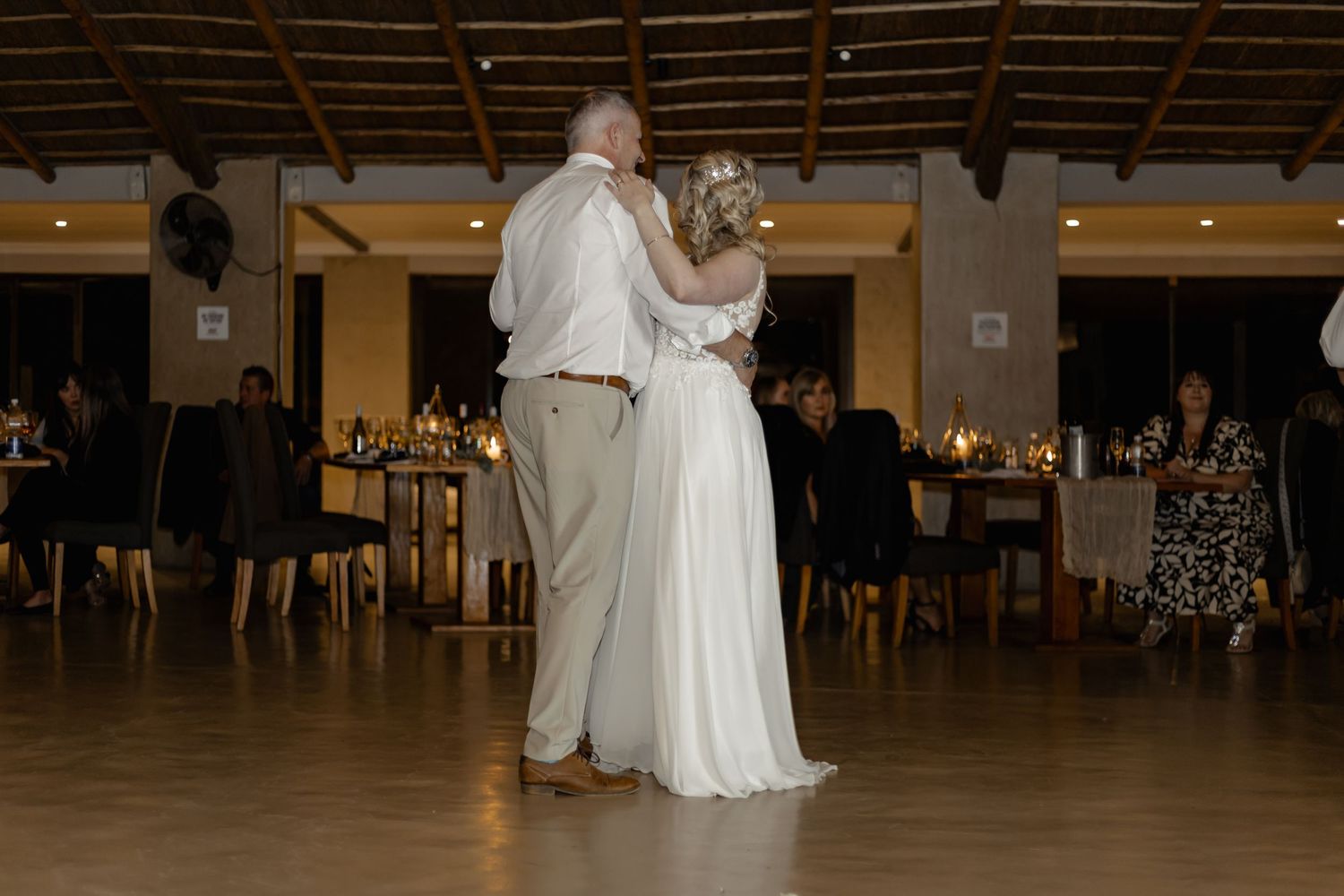 A couple shares their first dance as newlyweds in an elegantly decorated reception venue with wooden beams and warm lighting.