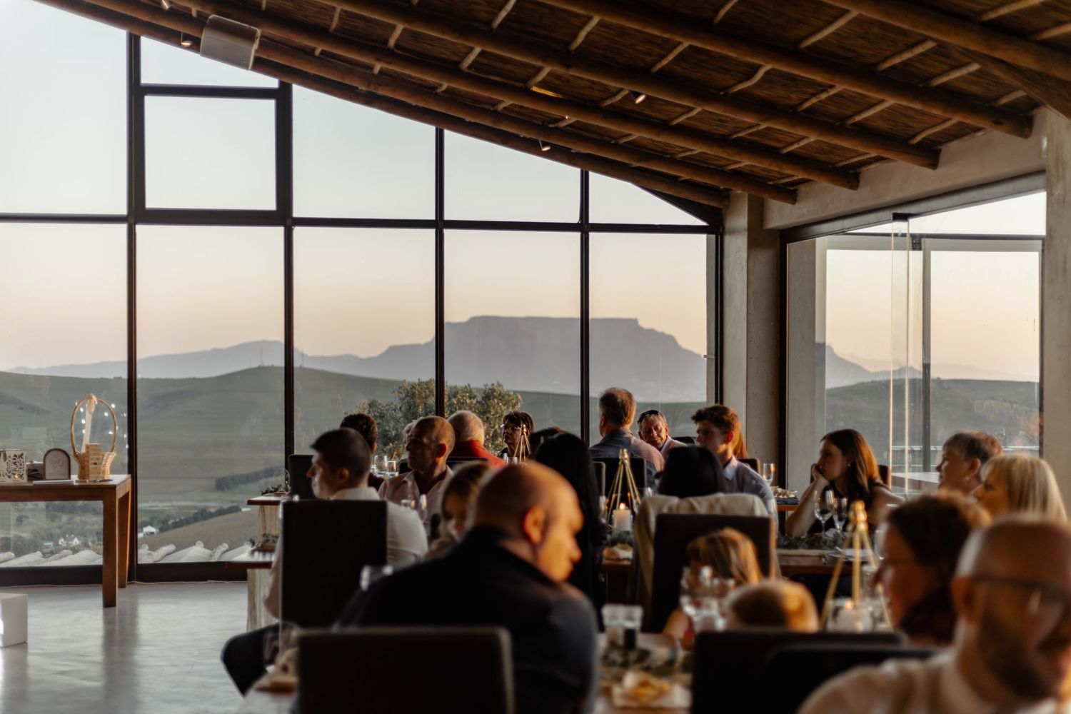 People dining at an upscale restaurant with panoramic mountain views through large windows at sunset.