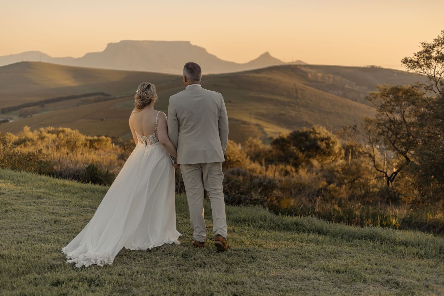 A couple in wedding attire sit on a hilltop overlooking mountains during golden hour sunset.