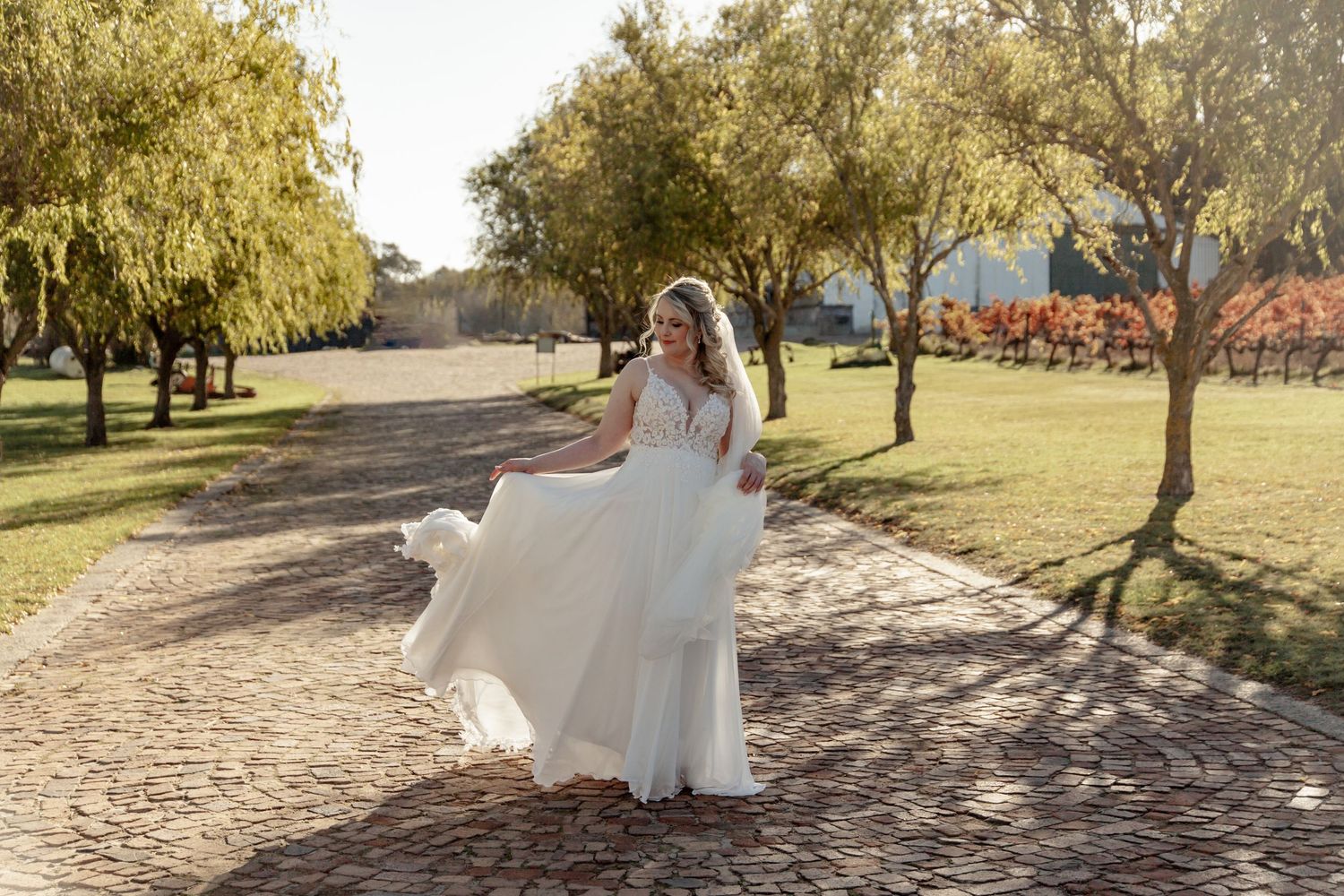 A person in a flowing white wedding dress spins gracefully on a sunlit path lined with autumn trees.