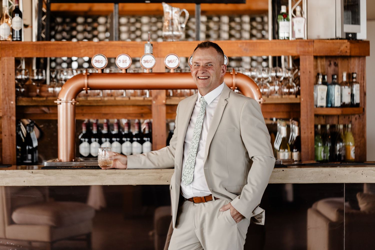 A person in a light colored suit leans casually against a bar counter with wine glasses and bottles displayed behind them.