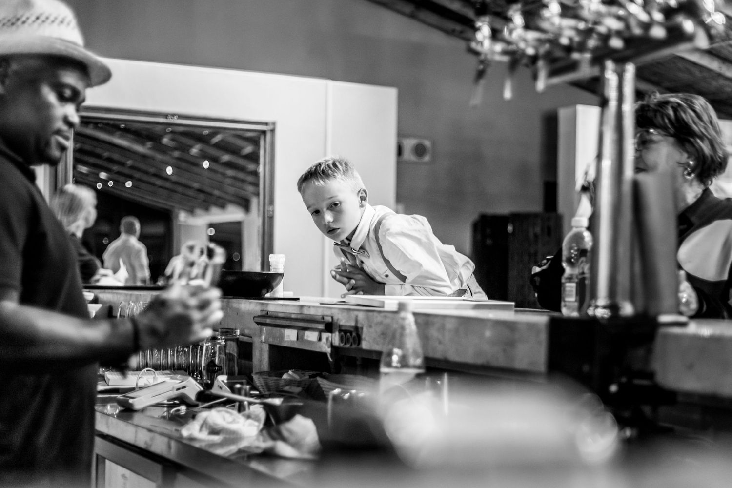 A black and white photograph shows a bartender interacting with patrons at a bar counter.
