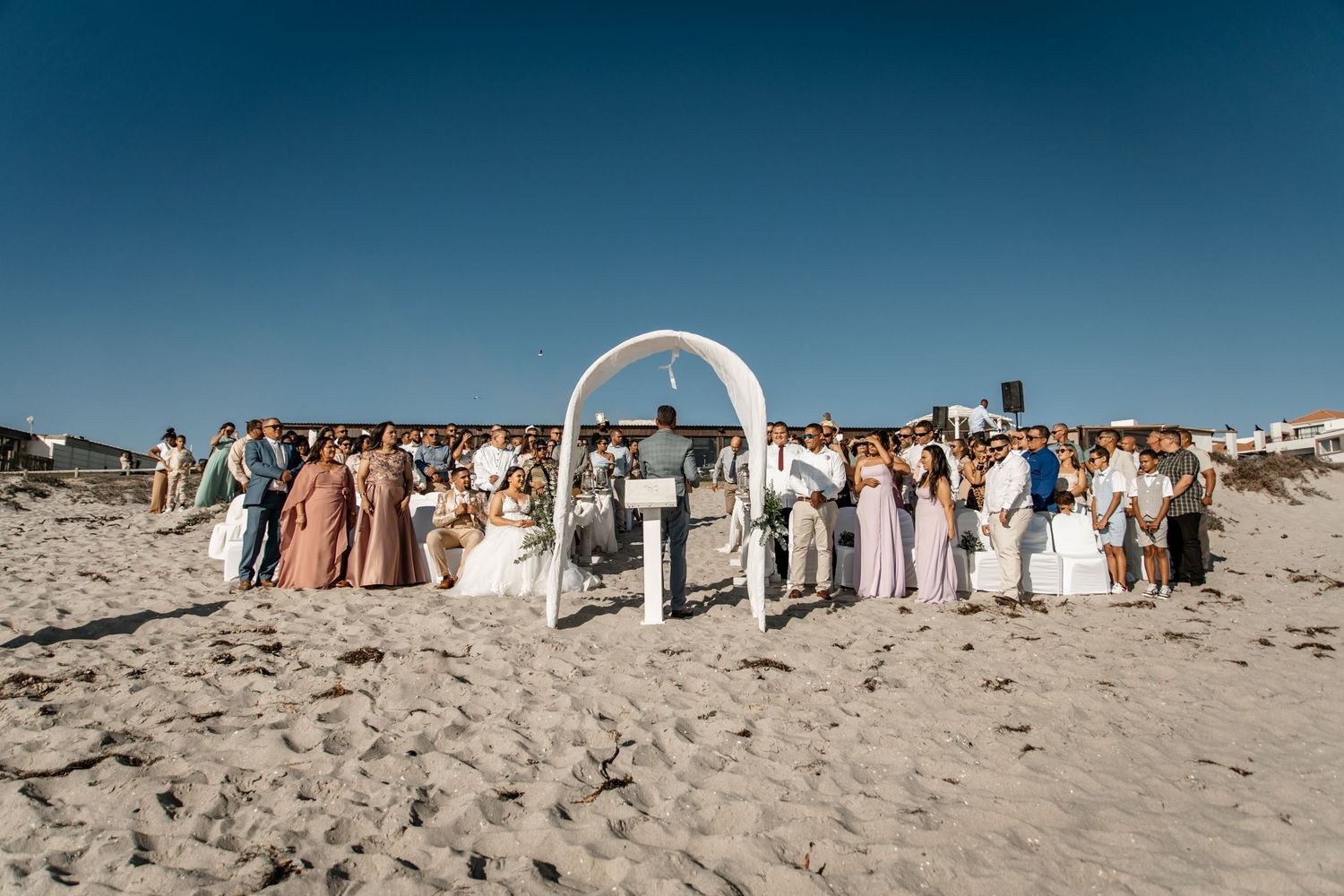 A beachside wedding ceremony under a white arch with guests gathered on sandy shores against a blue sky backdrop.
